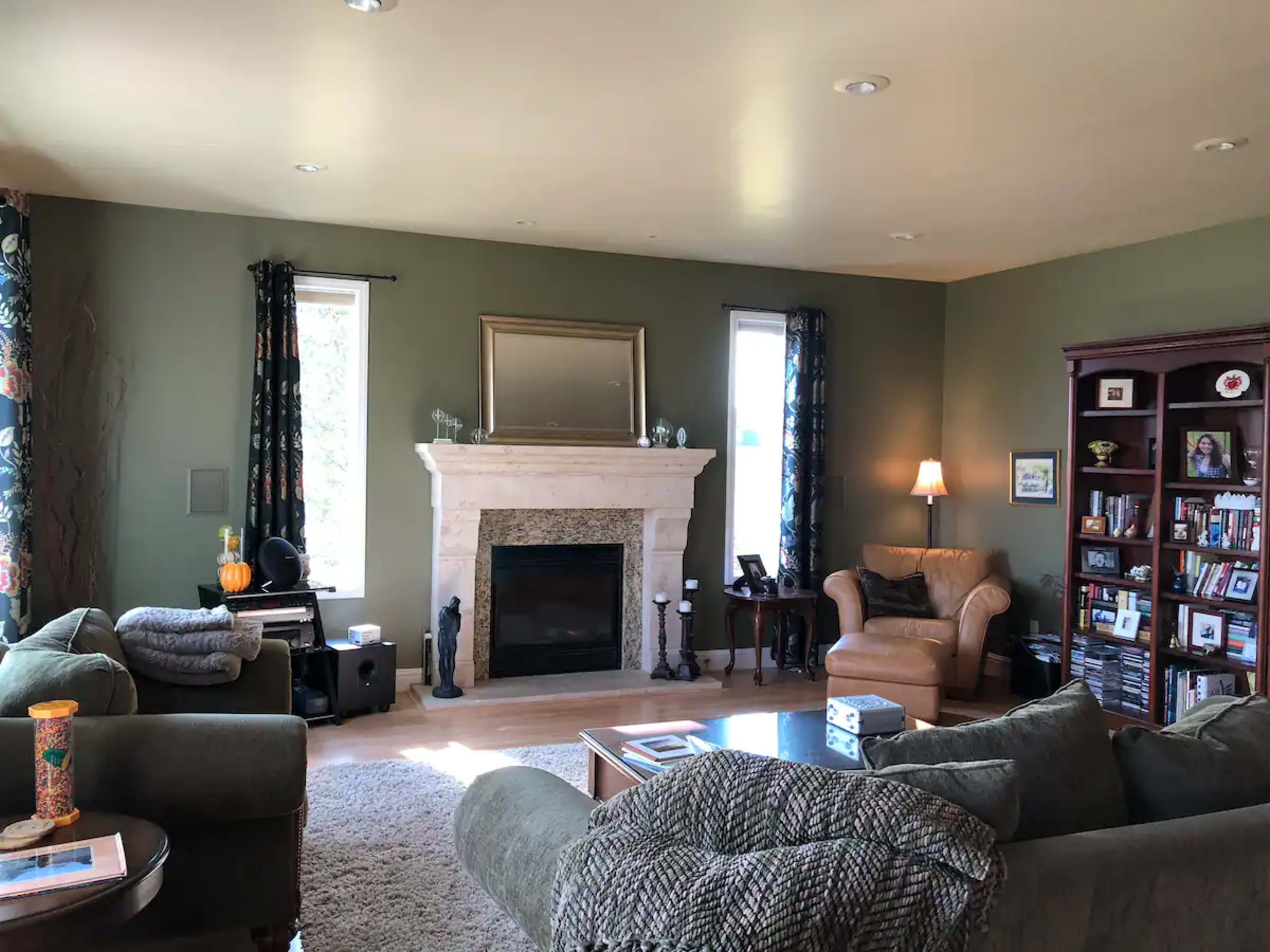 The image shows a living room with a fireplace, two windows, a bookshelf, and various pieces of seating furniture arranged around a coffee table.