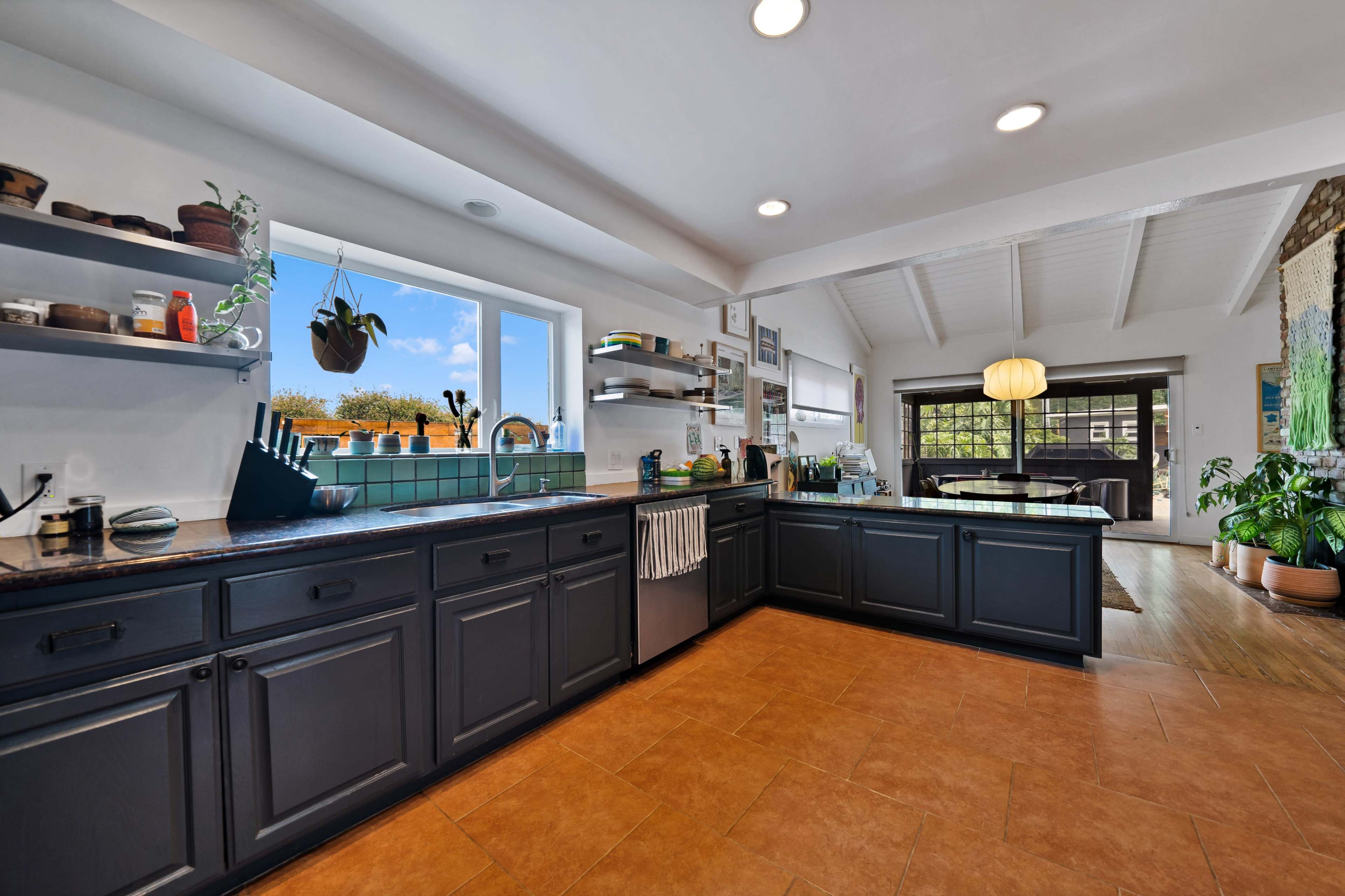 The kitchen features dark cabinets, a light countertop, and large windows that provide natural light, with a dining area visible in the background.