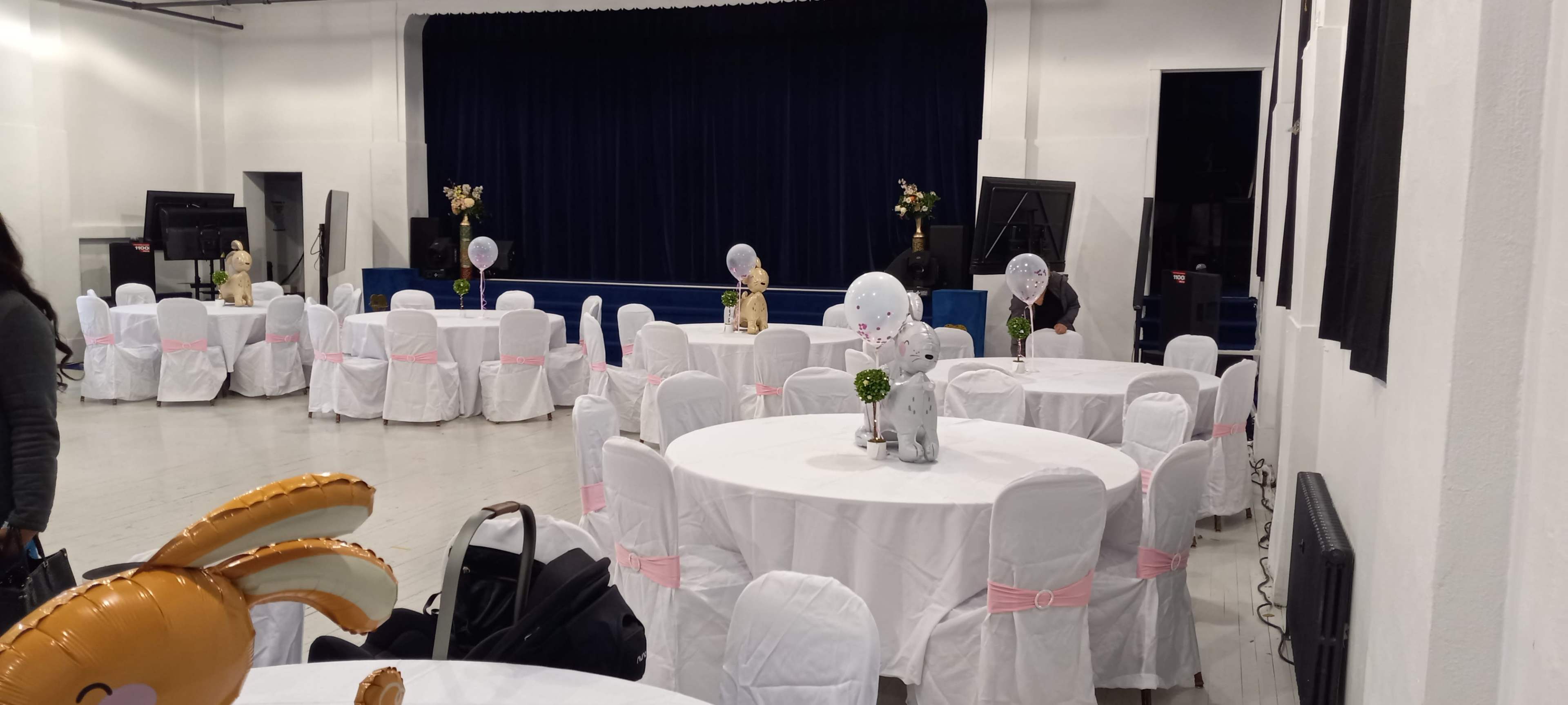 The image shows a banquet hall with round tables covered in white tablecloths and pink sashes, set up for an event, with a stage and decorations in the background.