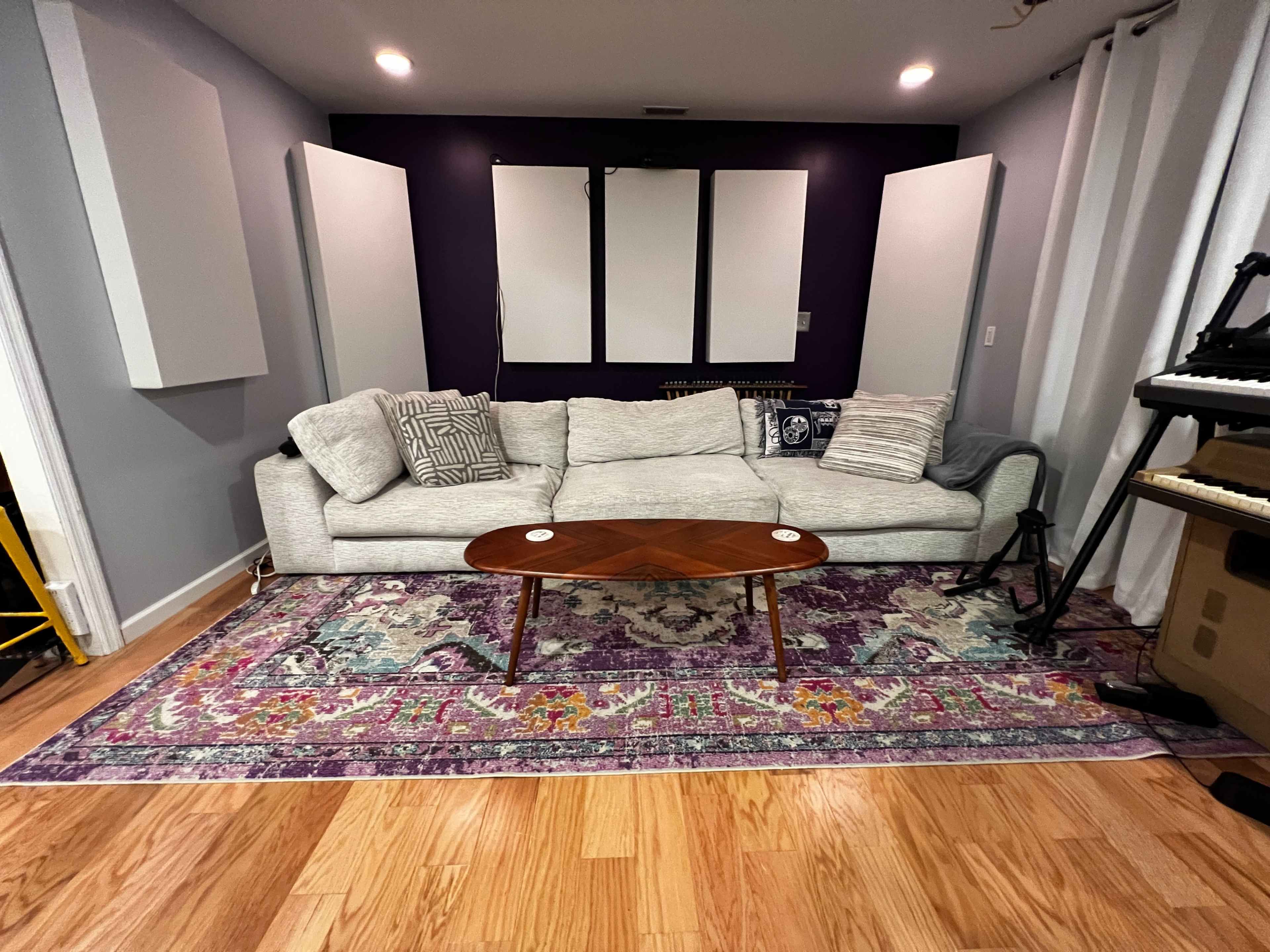 The image shows a living room with a gray sofa placed on a colorful area rug, flanked by soundproofing panels and a keyboard in the corner.