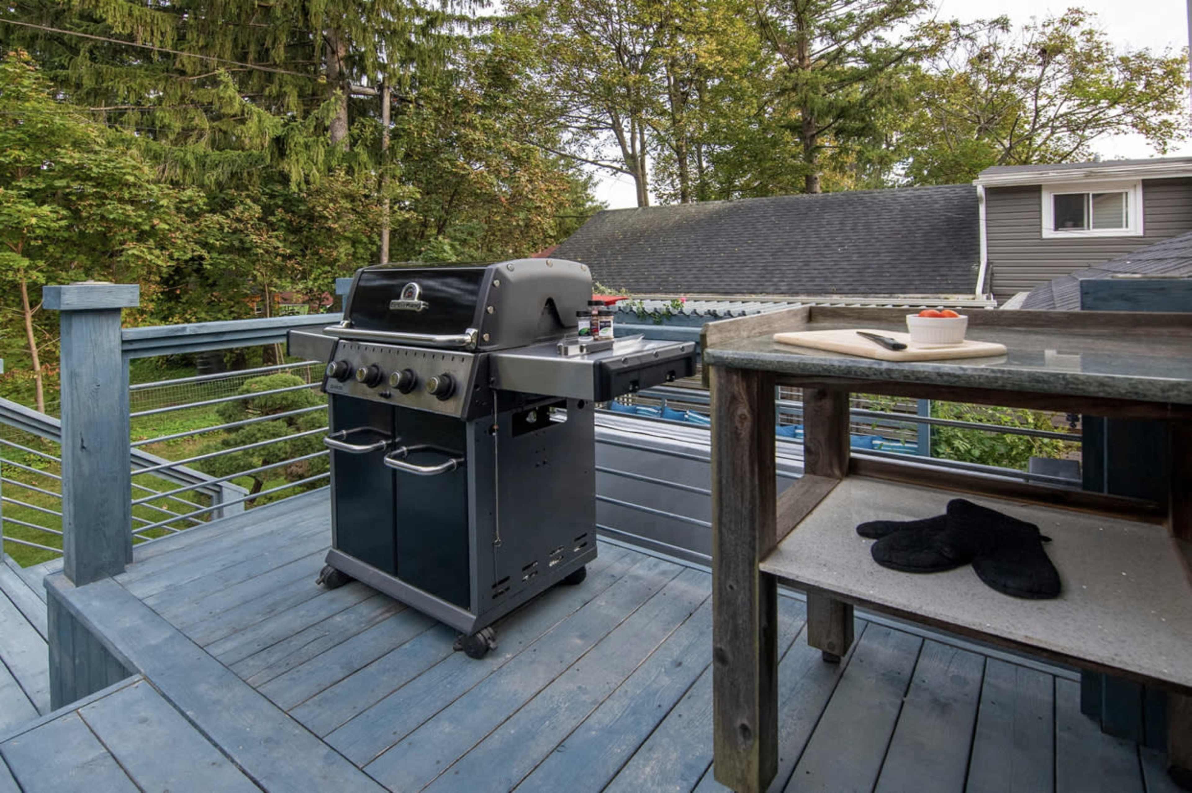 A black gas grill is positioned on a wooden deck with a table and a pair of slippers nearby, surrounded by trees and residential houses.
