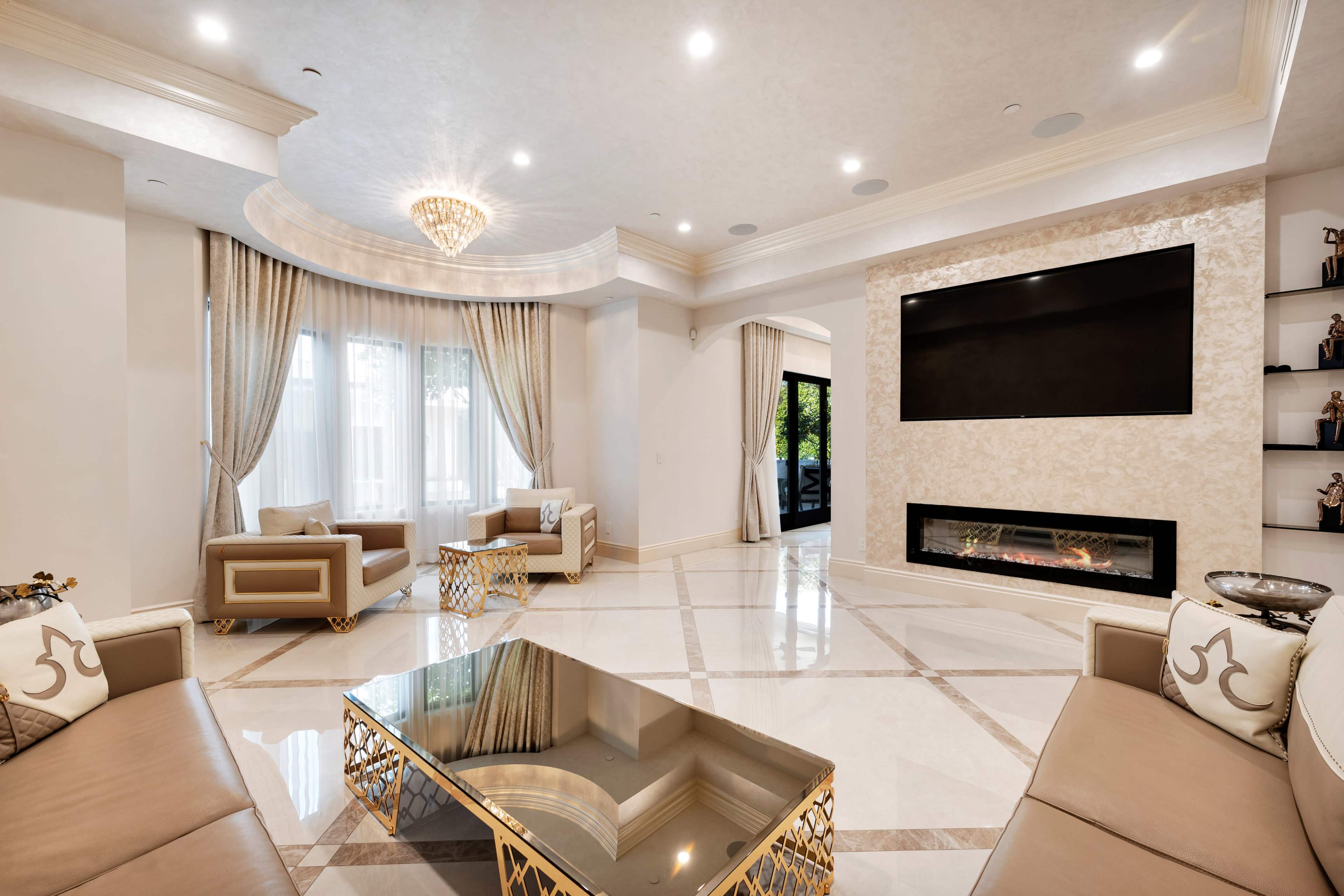 The image shows a spacious living room with marble flooring, elegant furniture, and a large flat-screen television mounted on the wall above a modern fireplace.
