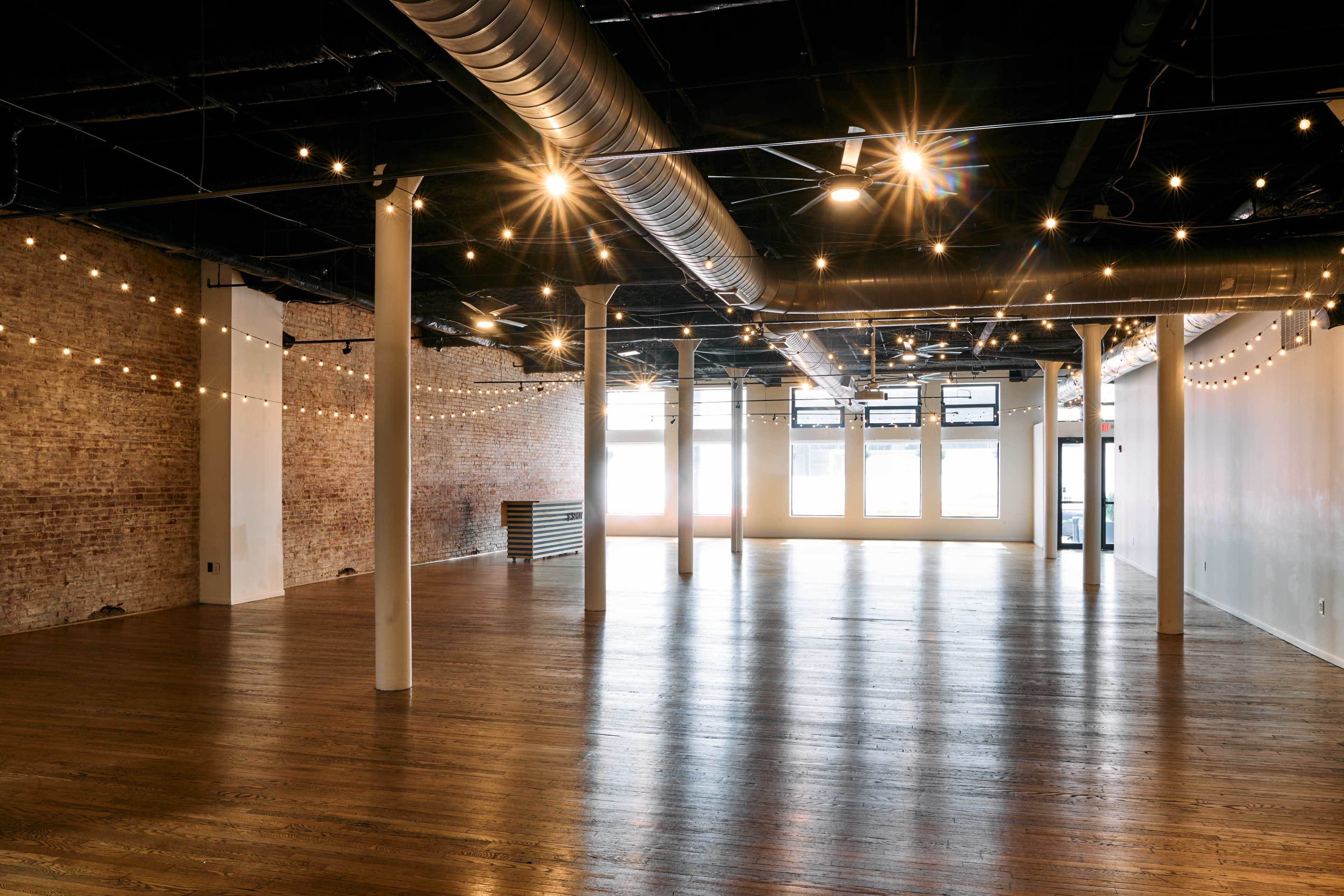 The image shows a spacious, empty room with wooden floors and exposed brick walls, illuminated by overhead string lights.
