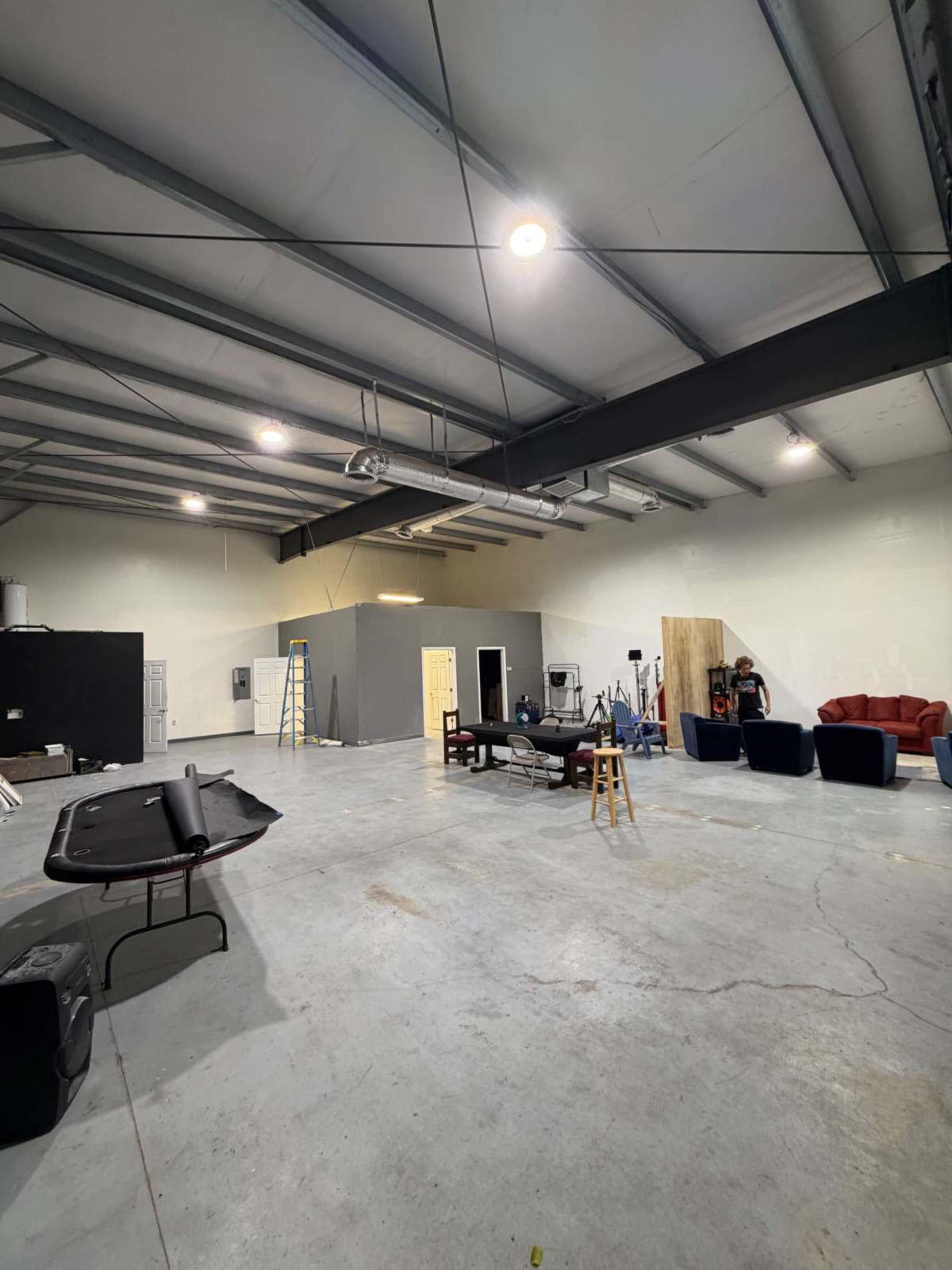 The image shows an industrial-style studio space with concrete floors, metal beams, and various pieces of furniture, including a couch and chairs, arranged in the center.