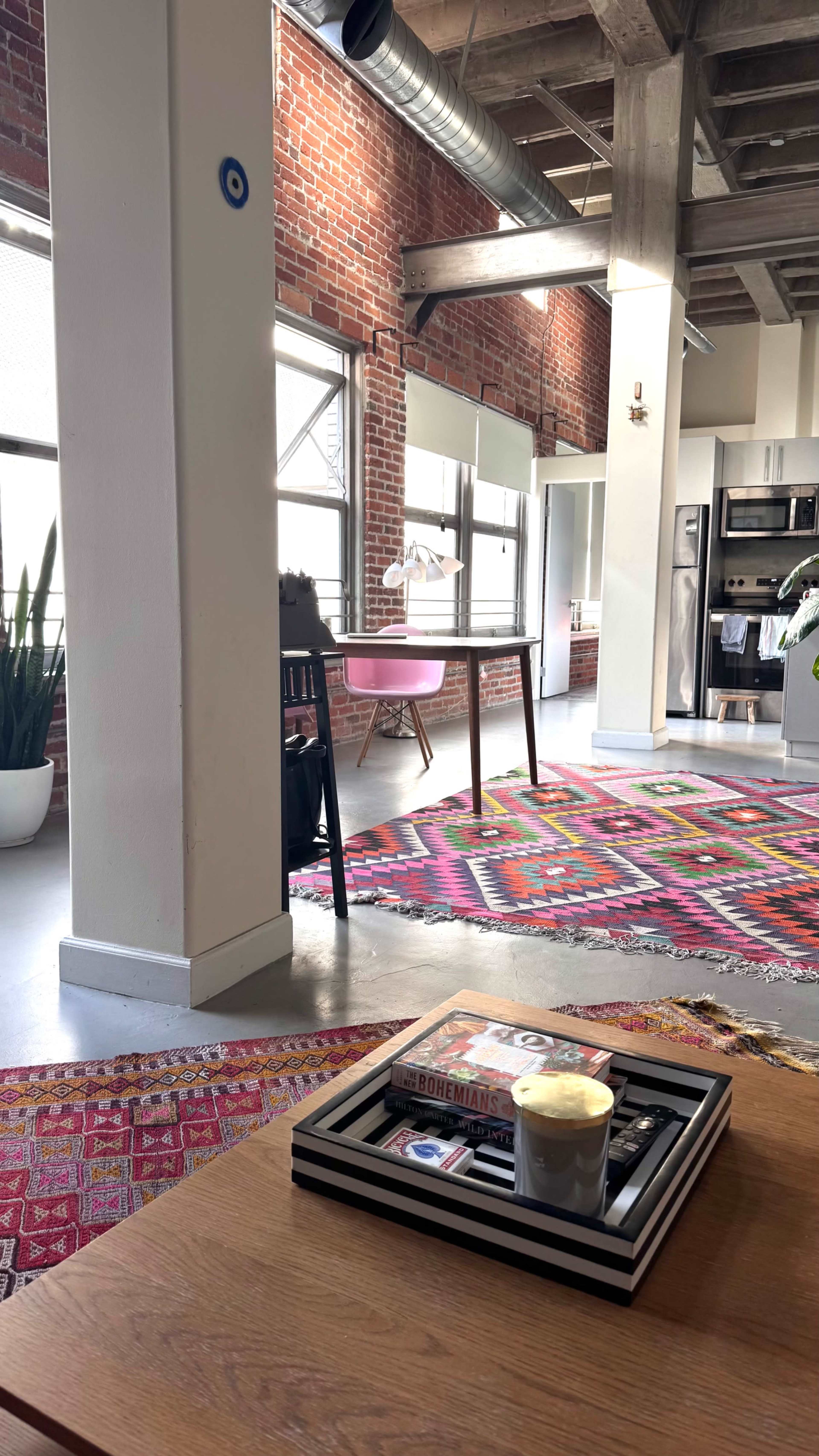 The image depicts a modern, industrial-style living space featuring a pink desk, patterned area rug, and exposed brick walls.