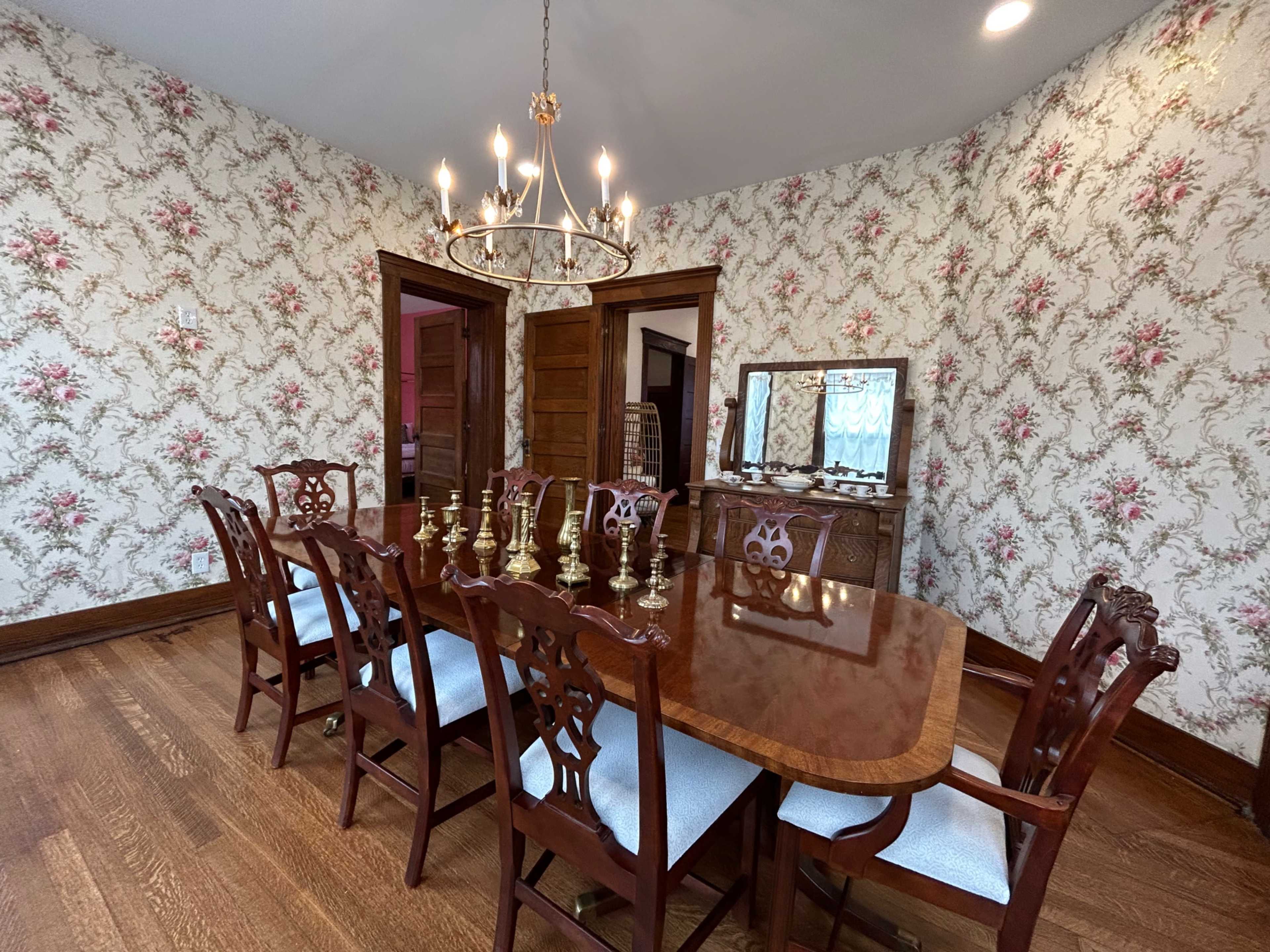 A dining room with a large wooden table surrounded by eight ornate chairs, a chandelier above, and floral wallpaper.