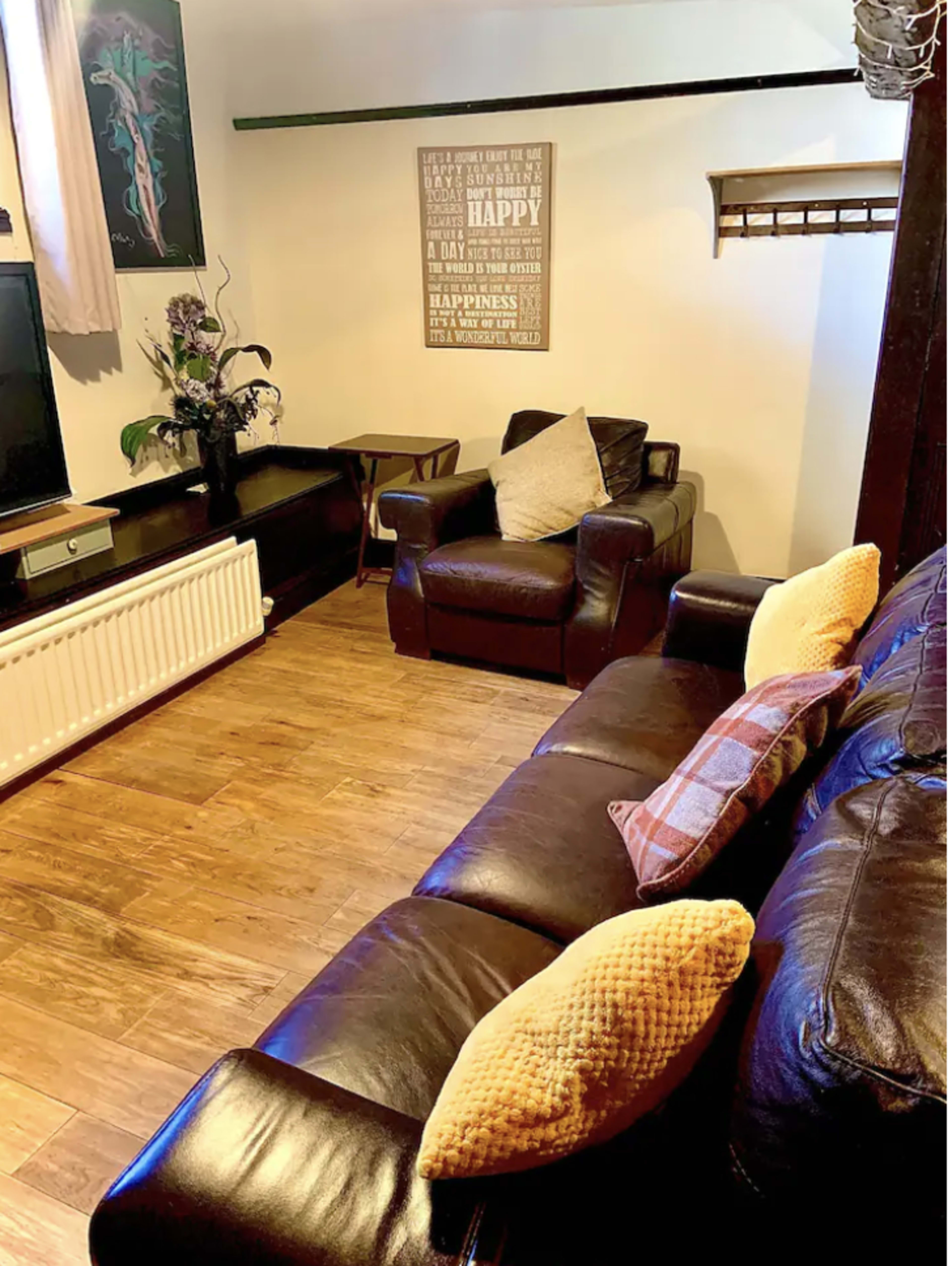 The image shows a living room with a leather sofa and armchair, a television unit, a decorative wall plaque, and a wooden floor.