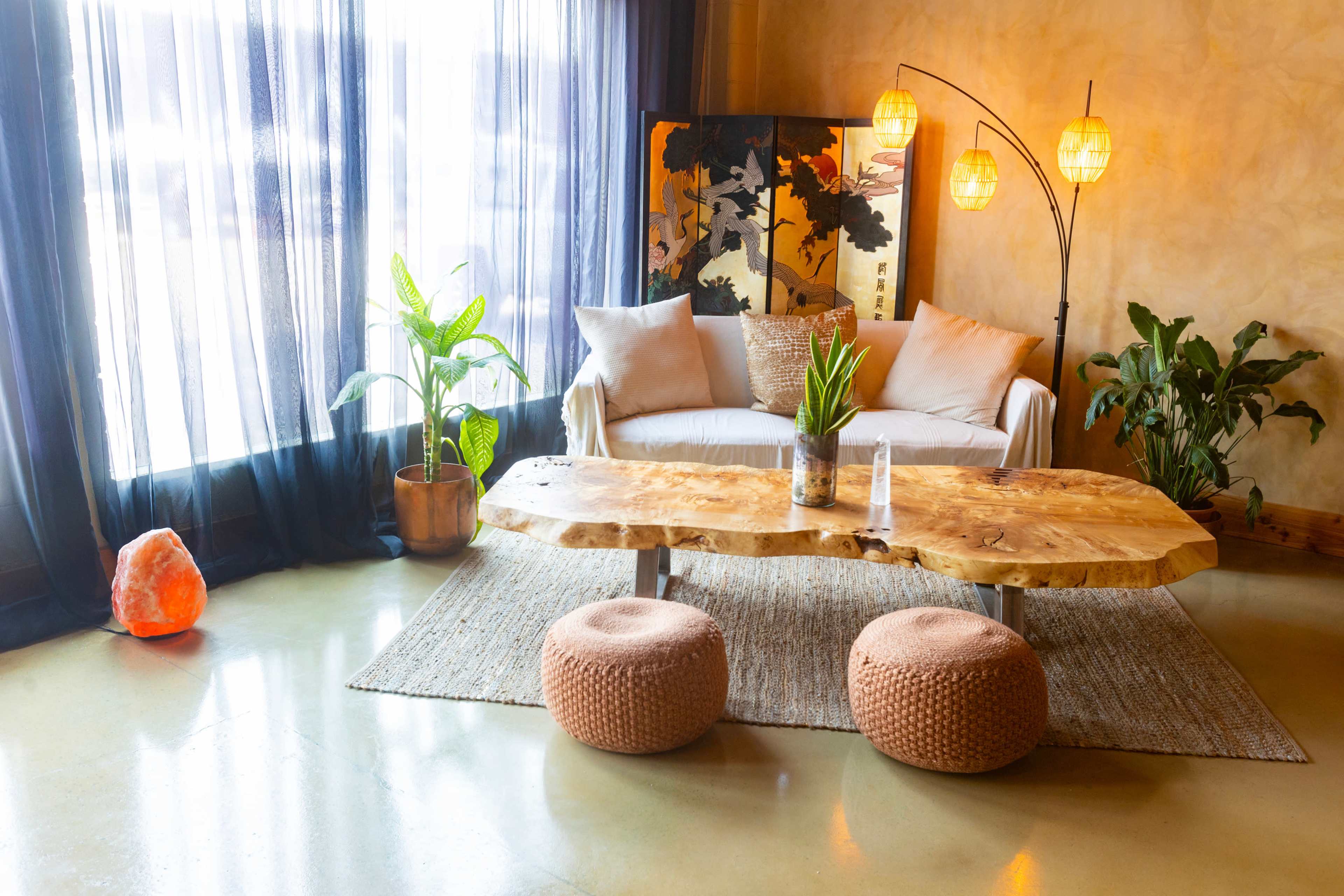 The scene depicts a modern living room with a white sofa, a large wooden coffee table, and various plants, illuminated by natural light filtering through sheer curtains.