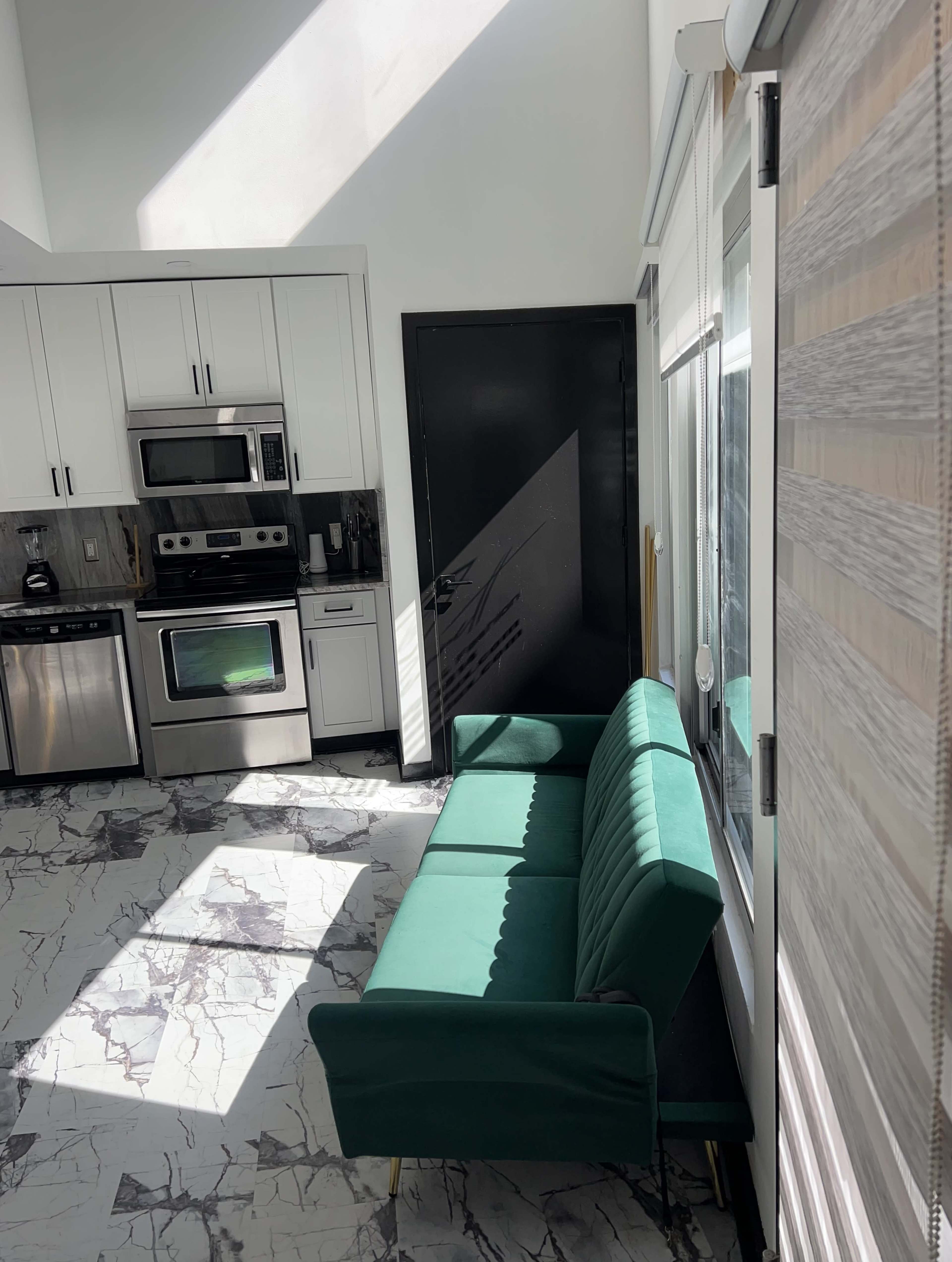 The image depicts a modern kitchen with white cabinets, stainless steel appliances, and a green velvet sofa positioned near a large window.