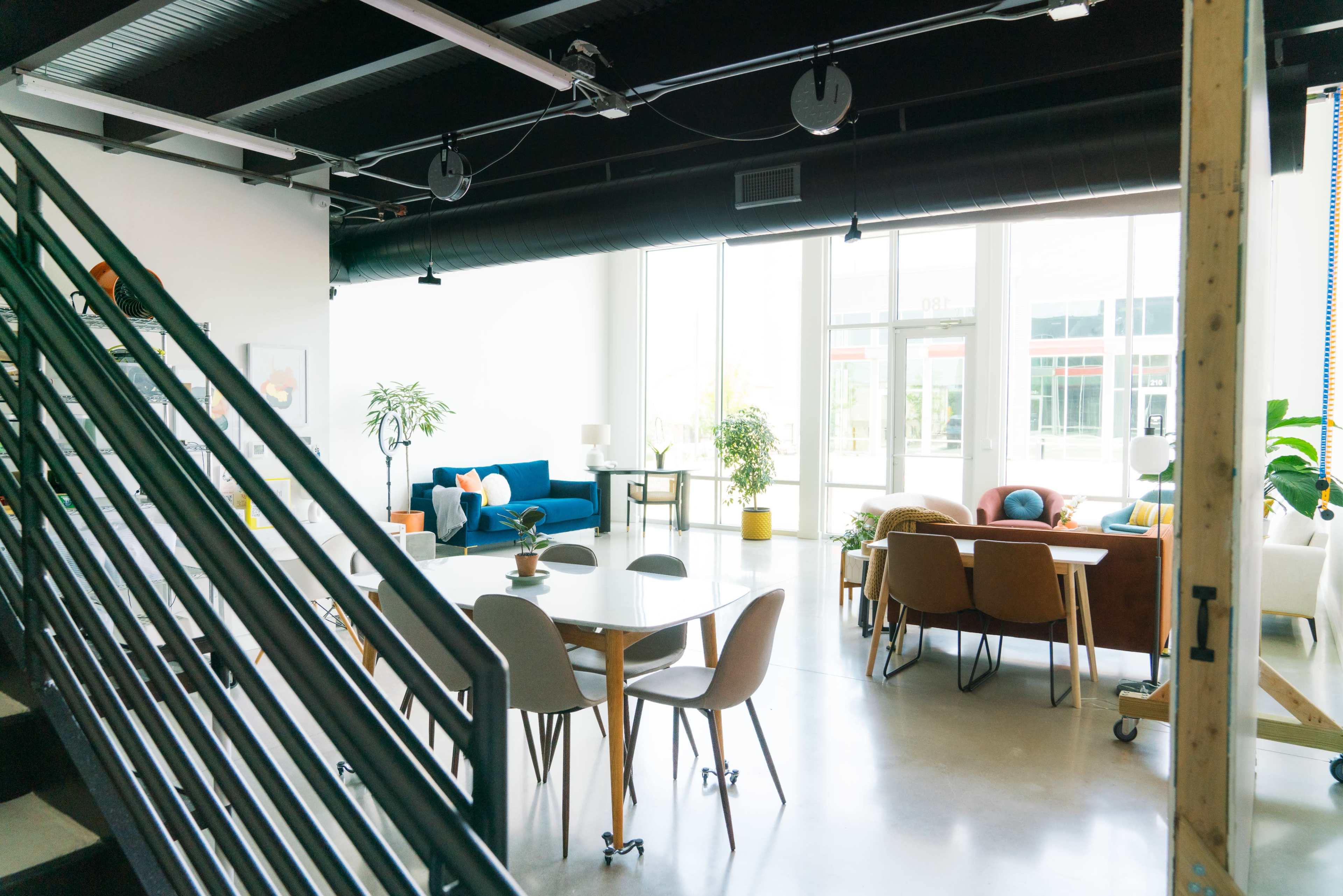 A modern, open-plan workspace features a dining table surrounded by chairs, a staircase, and various seating areas with plants and natural light.