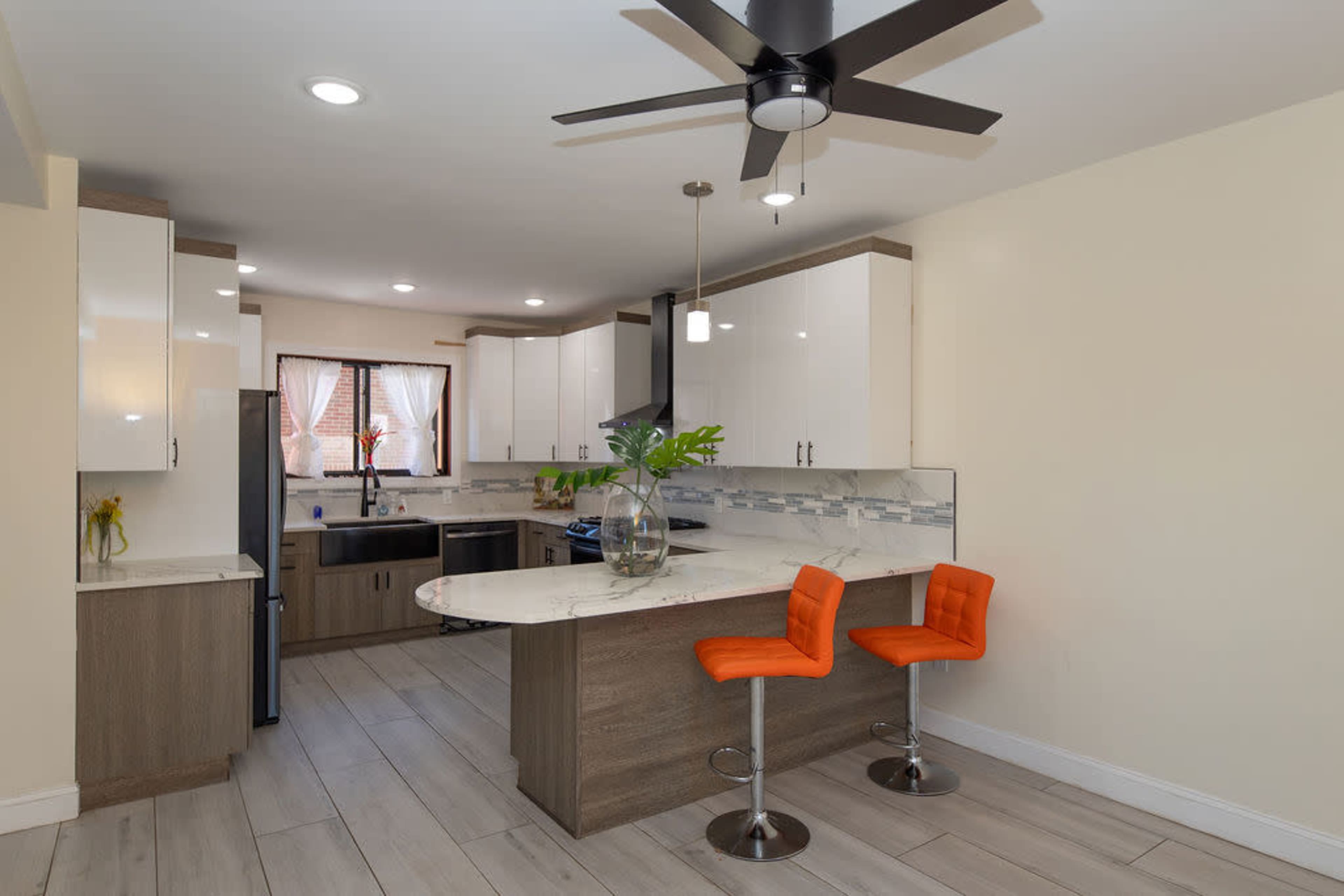 A modern kitchen with light-colored cabinets, a marble countertop, and two orange bar stools at a circular countertop.