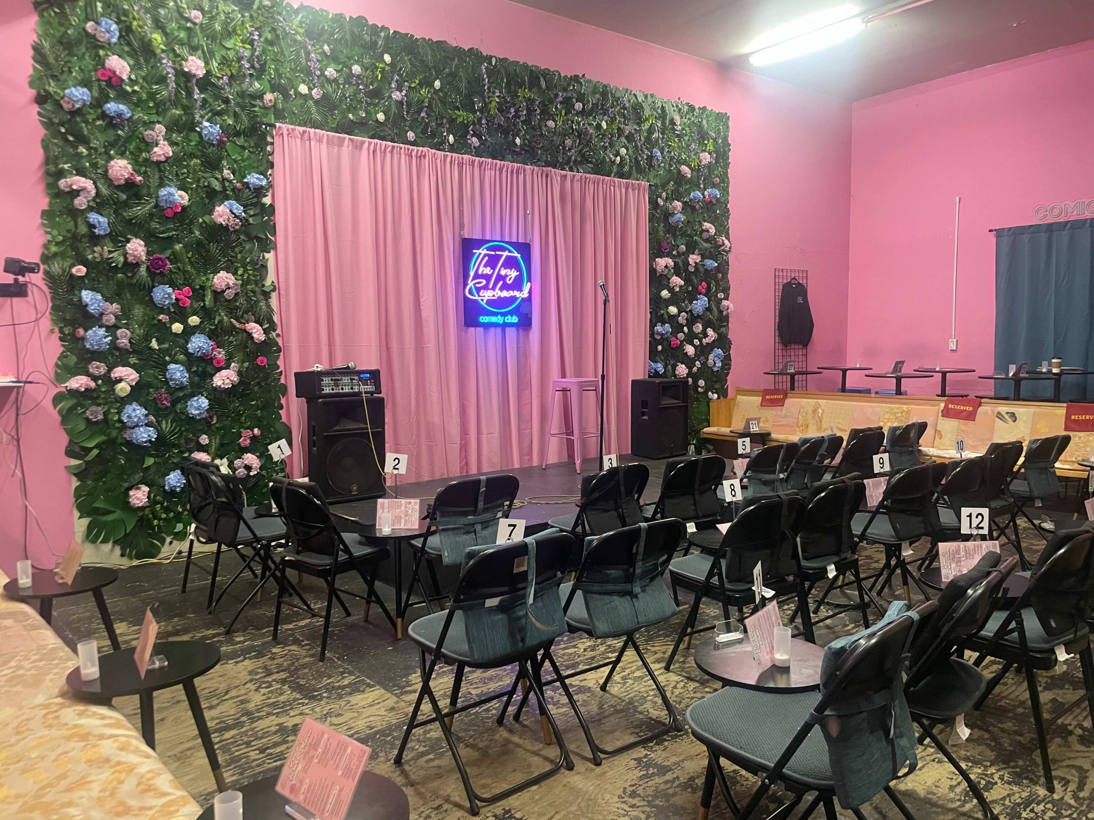 A small performance space with rows of black chairs arranged facing a pink curtain adorned with flowers and a neon sign, with a microphone stand in the center.