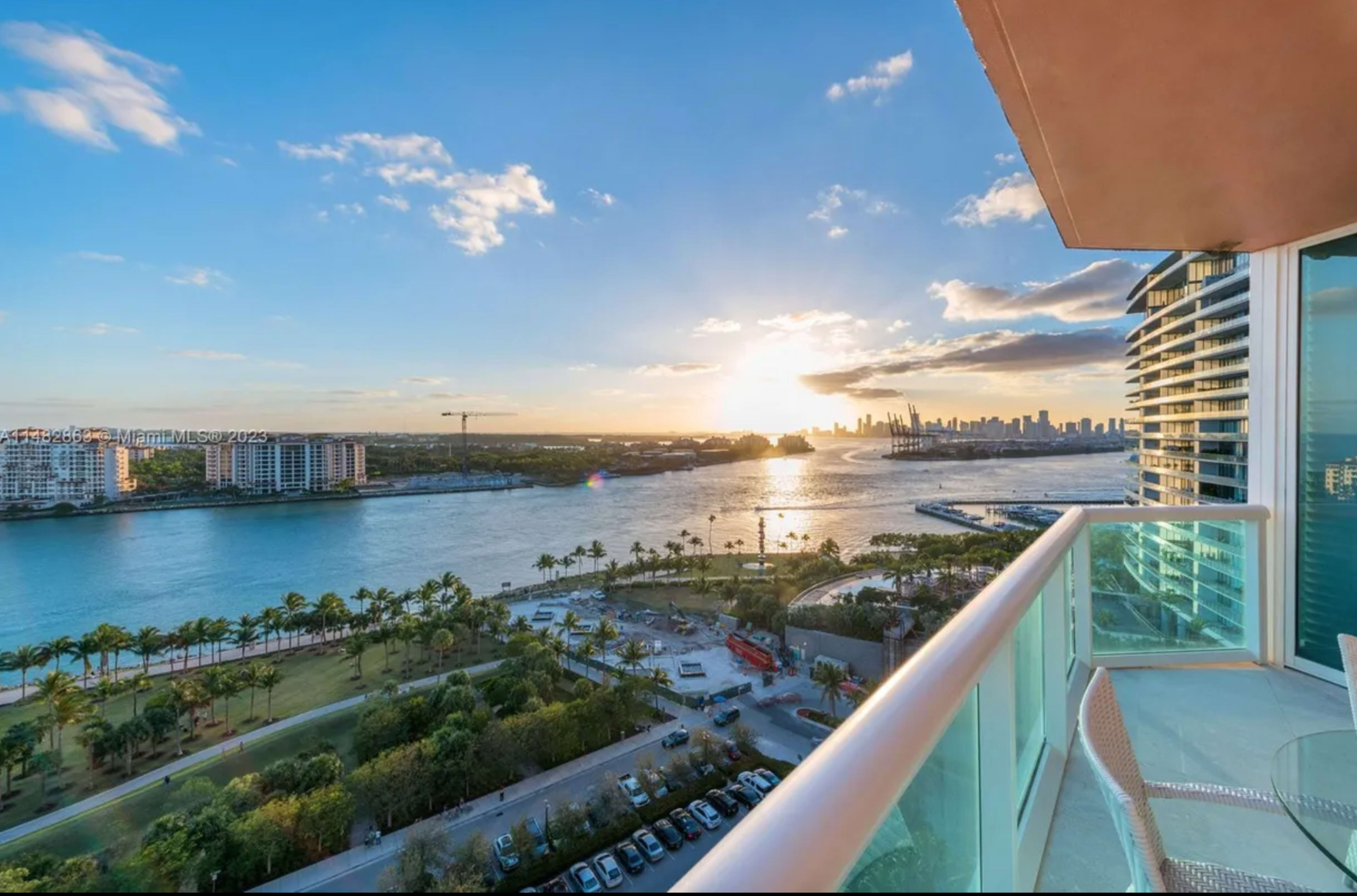 A balcony view overlooking a river at sunset, with a skyline in the distance and palm trees lining the waterfront.