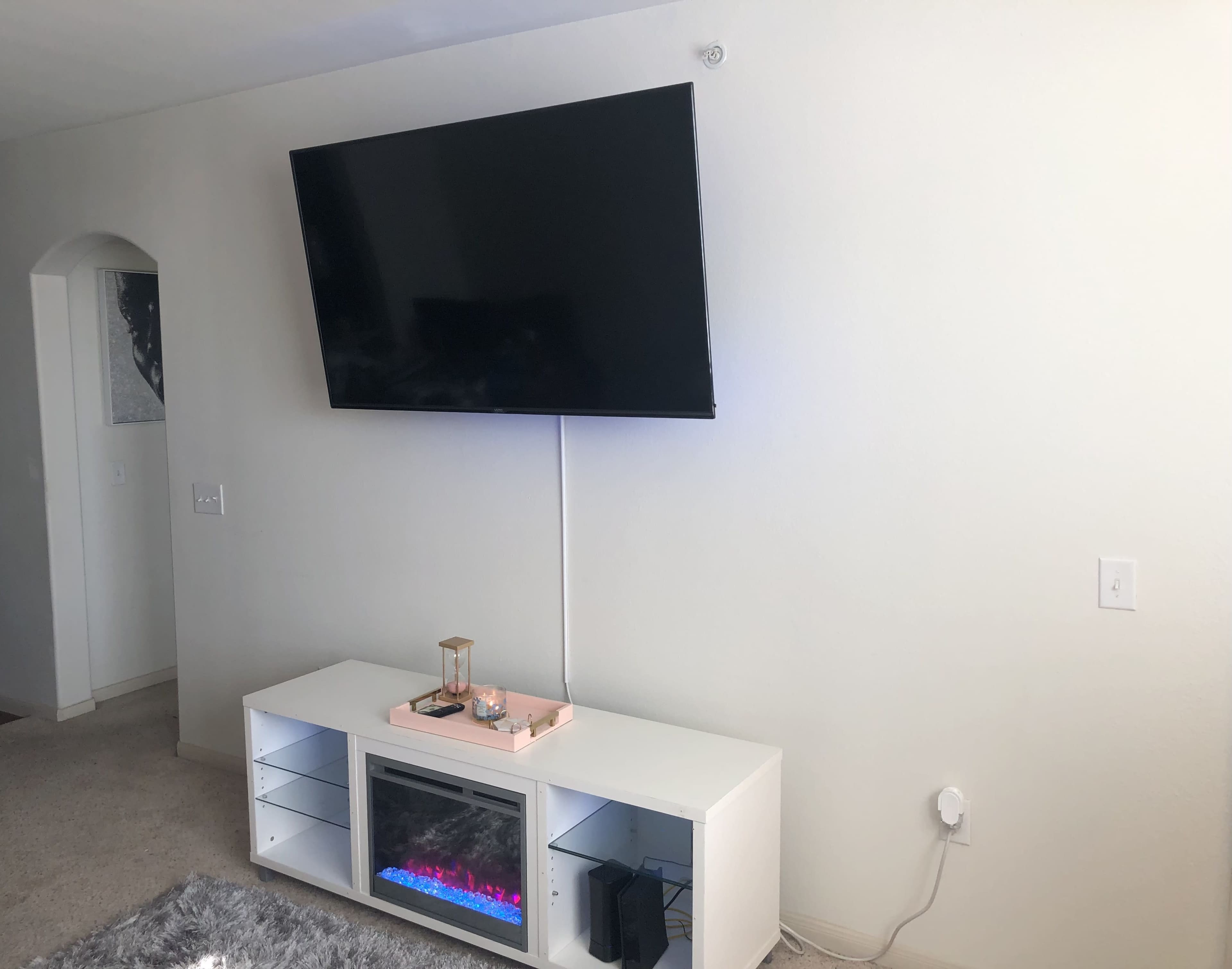 A wall-mounted flat-screen television above a white media console with an electric fireplace in the center and a decorative item on top.