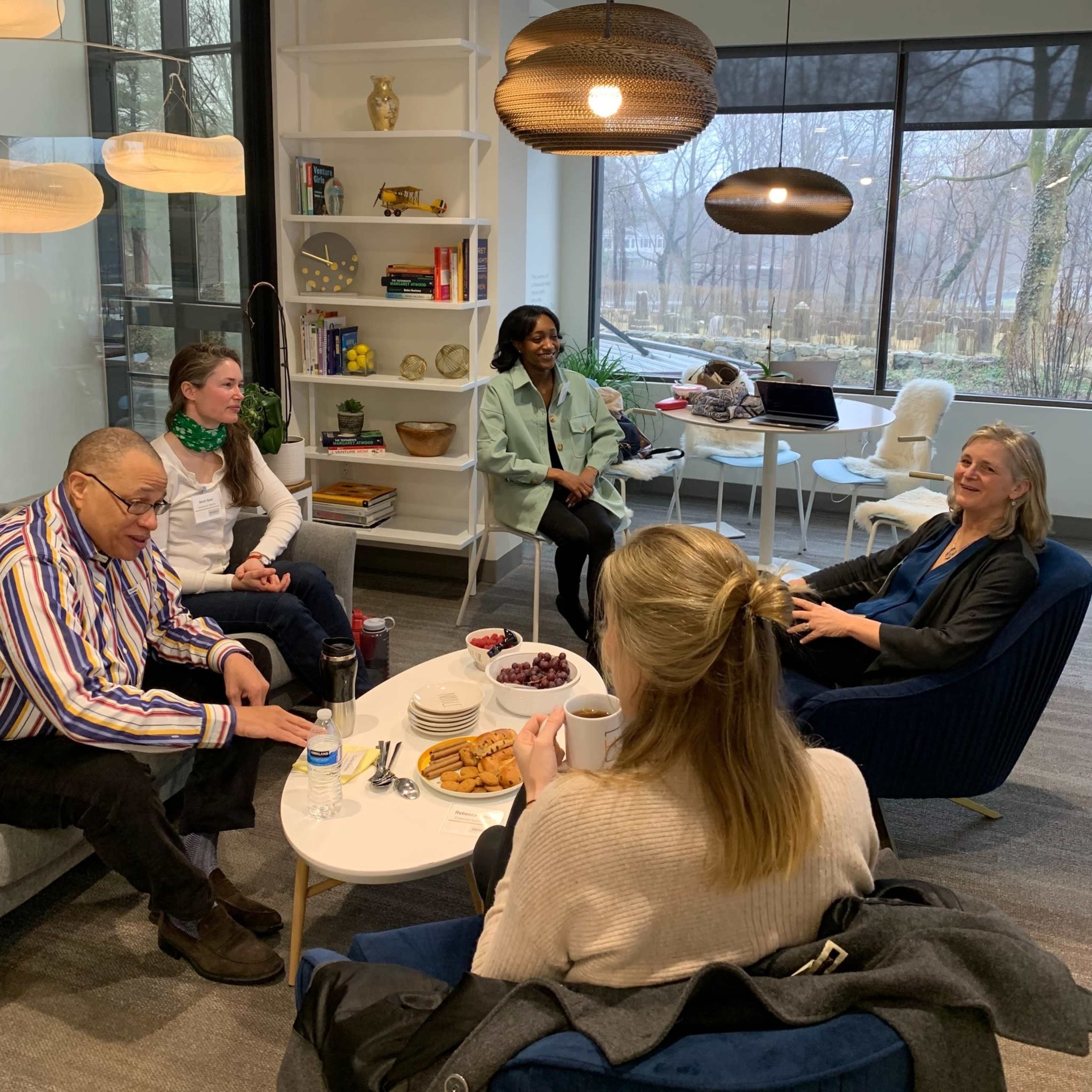 Five people are seated in a modern lounge area, engaged in conversation, with snacks and drinks on a coffee table.