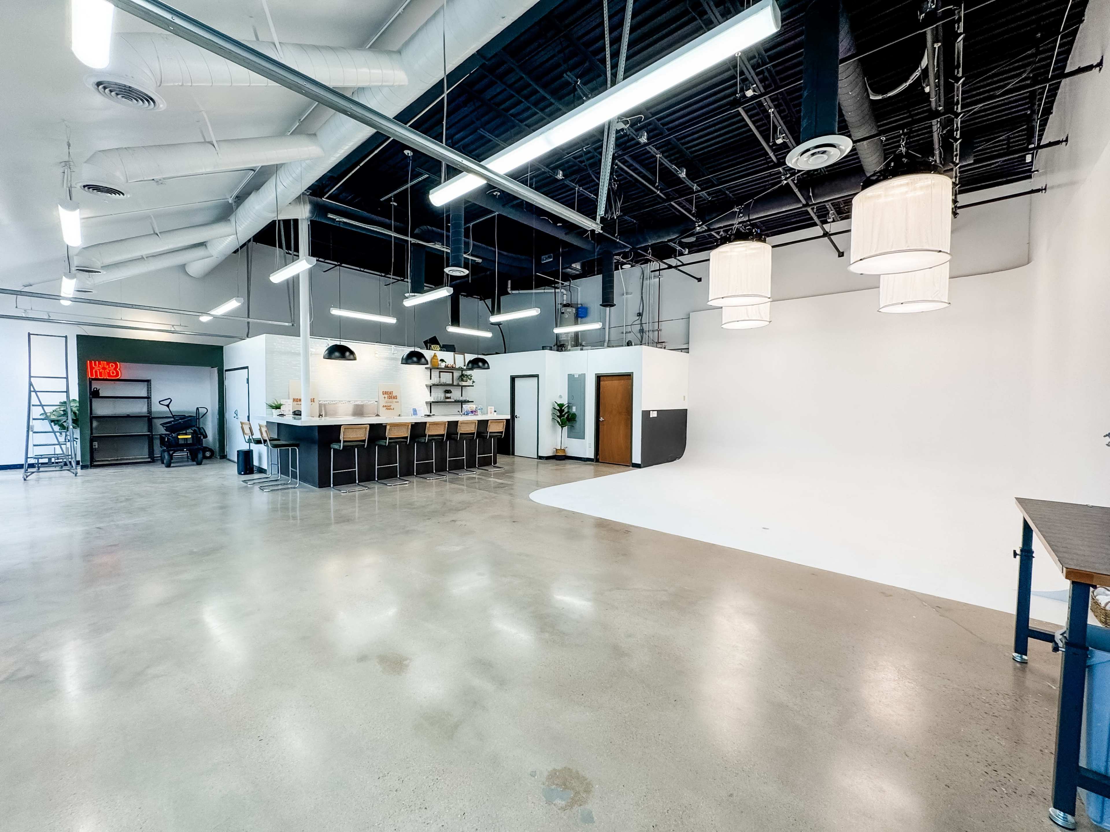The image shows a spacious, well-lit interior featuring a large open area with polished concrete floors, a kitchen counter, and a white backdrop.