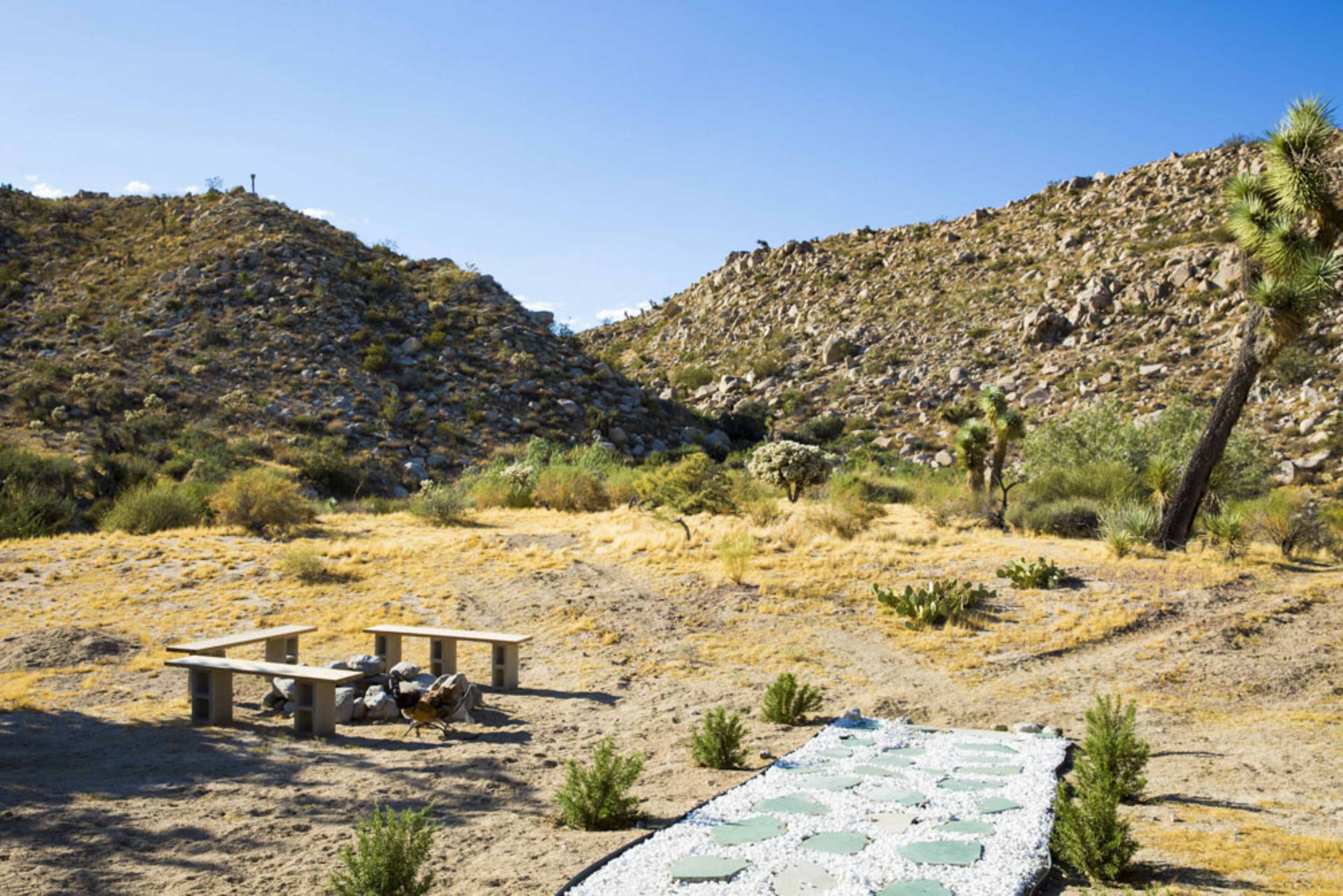 The image shows a dry desert landscape with a stone pathway leading to a simple wooden bench area surrounded by sparse vegetation and rocky hills.