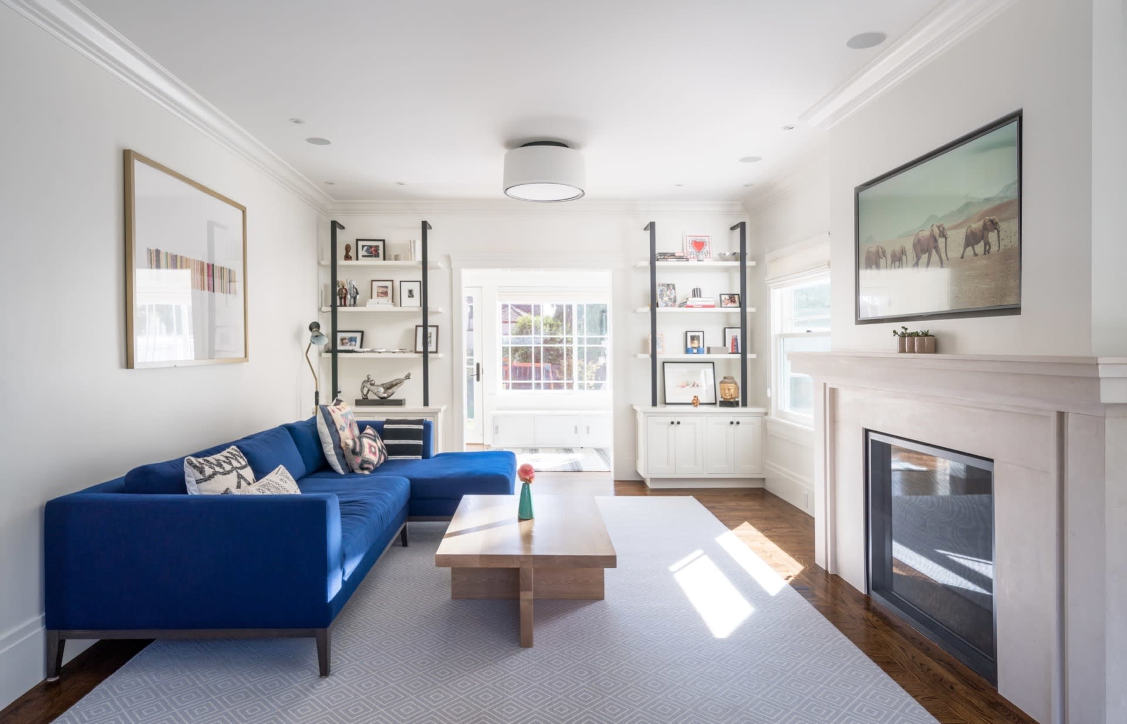 A modern living room features a blue sectional sofa, a wooden coffee table, built-in shelves, and a fireplace beneath a large window.