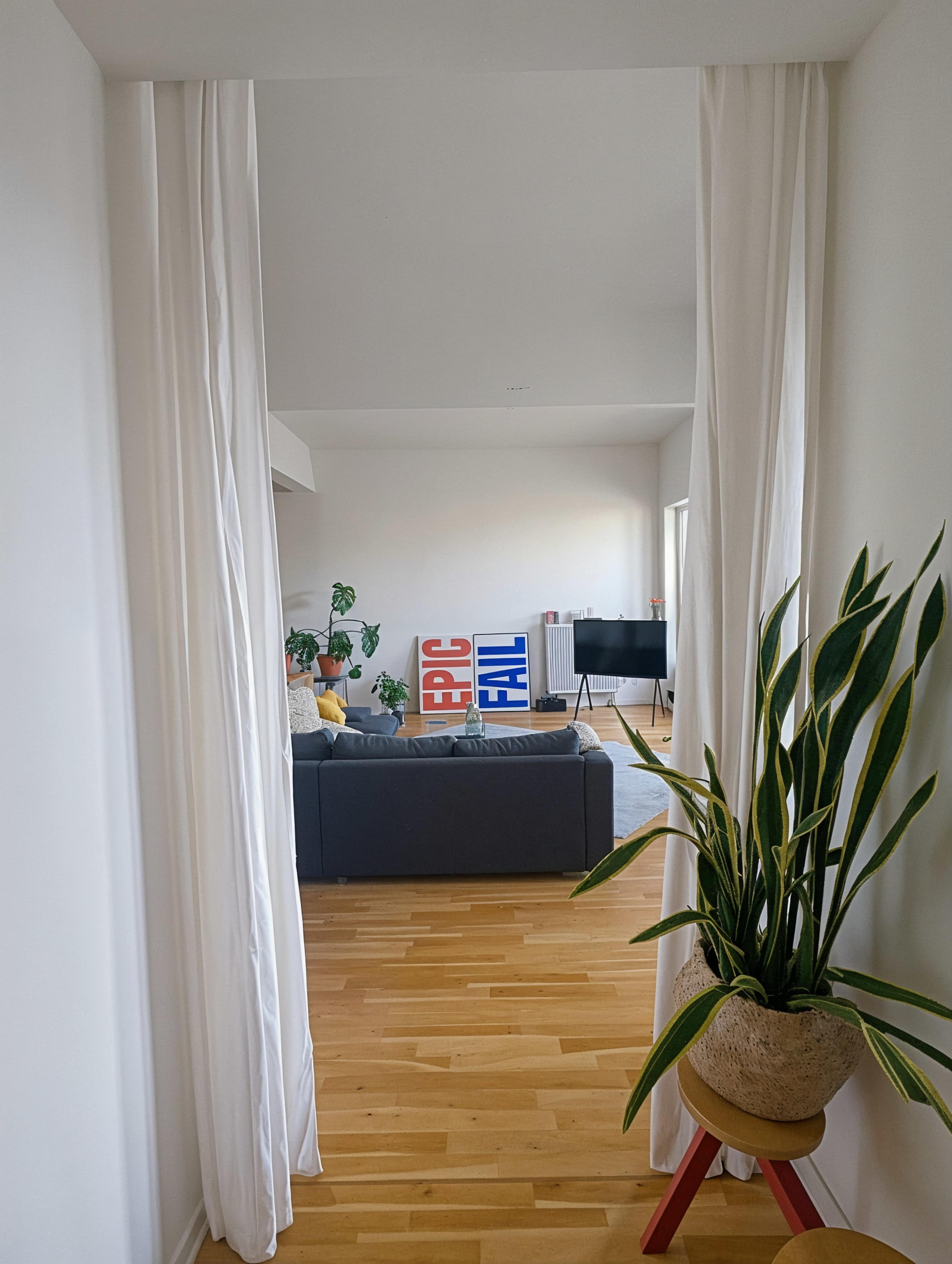 A hallway with white curtains leads to a living room featuring a gray sofa, a television, and decorative plants.
