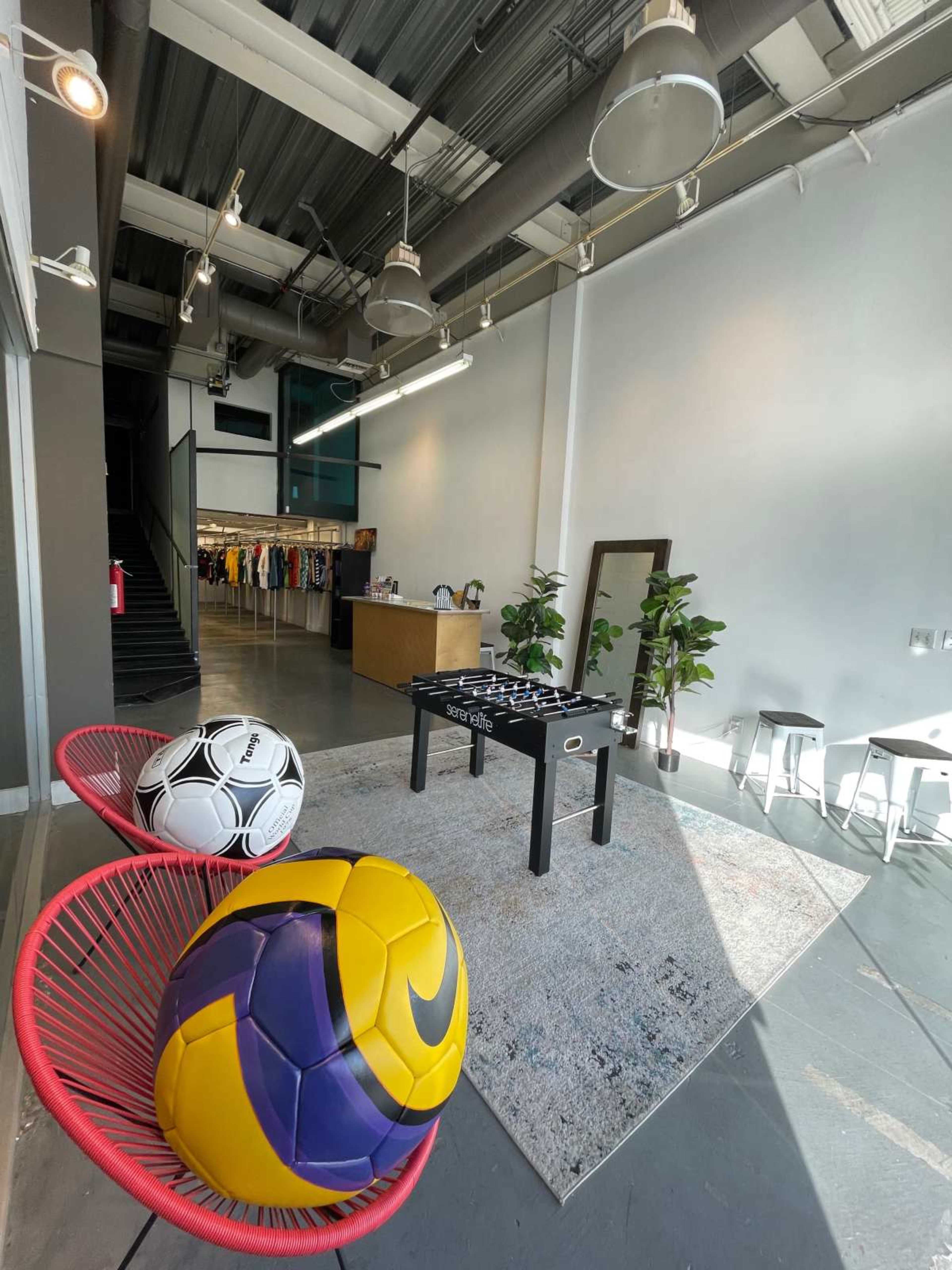 The image shows a modern retail space with colorful soccer balls on red chairs, a foosball table on a rug, and clothing displayed in the background.