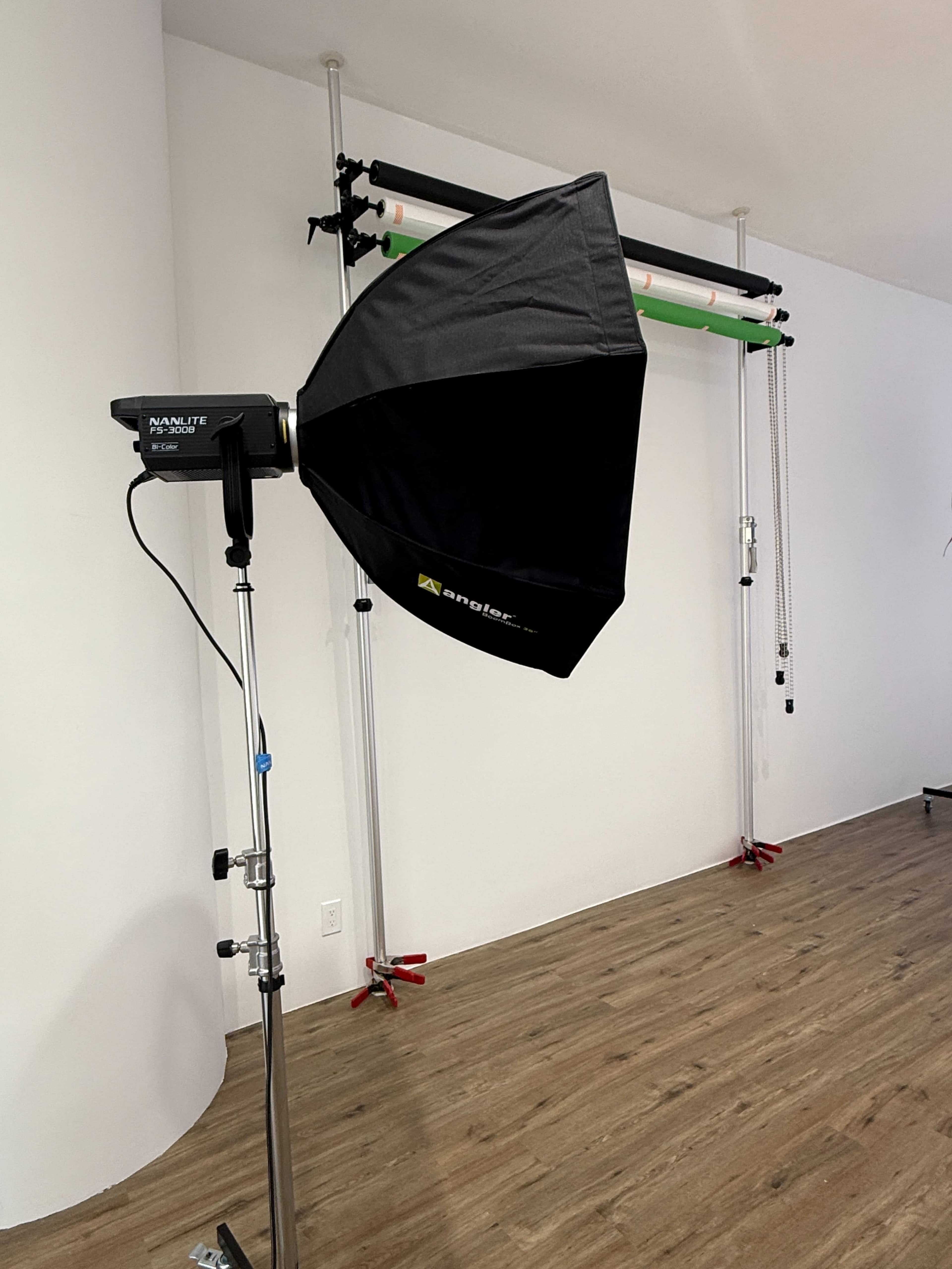A black softbox is mounted on a stand in a studio setting, with backdrop support poles in the background.