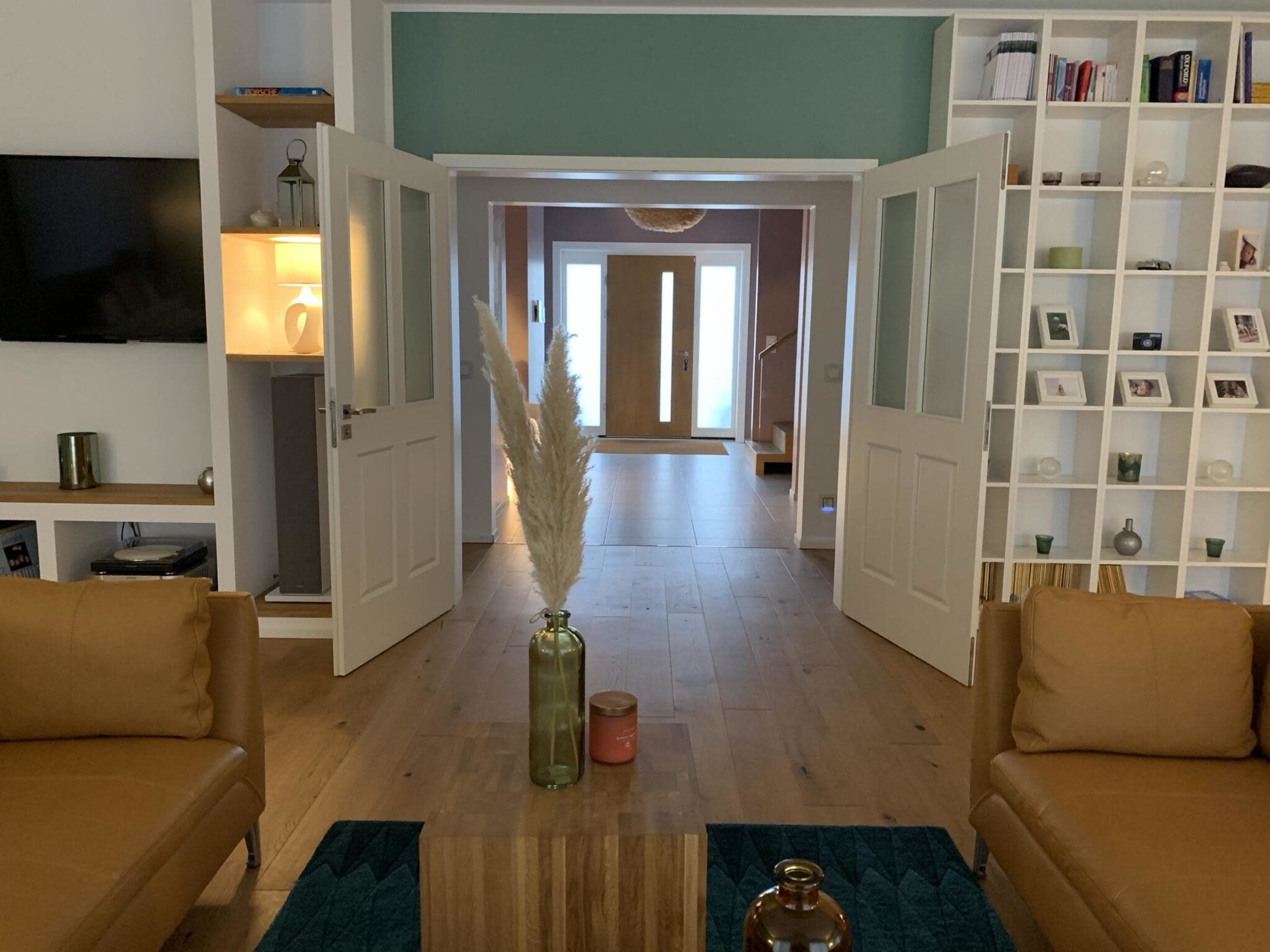A living room with two tan couches facing a coffee table, leading to a hallway with double doors and a light-colored entryway.