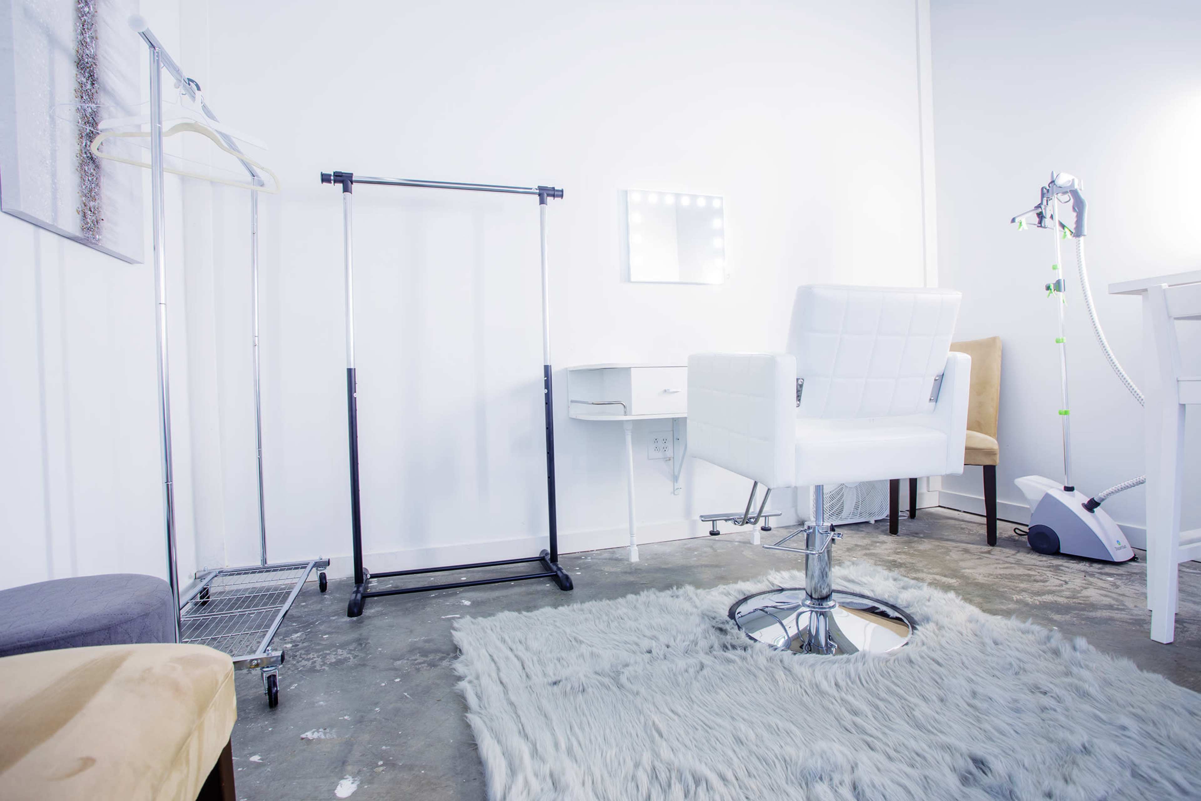 The image shows a minimalist salon space featuring a white chair on a circular chrome base, a clothing rack, a small table with a mirror, and a cosmetic device in the corner.