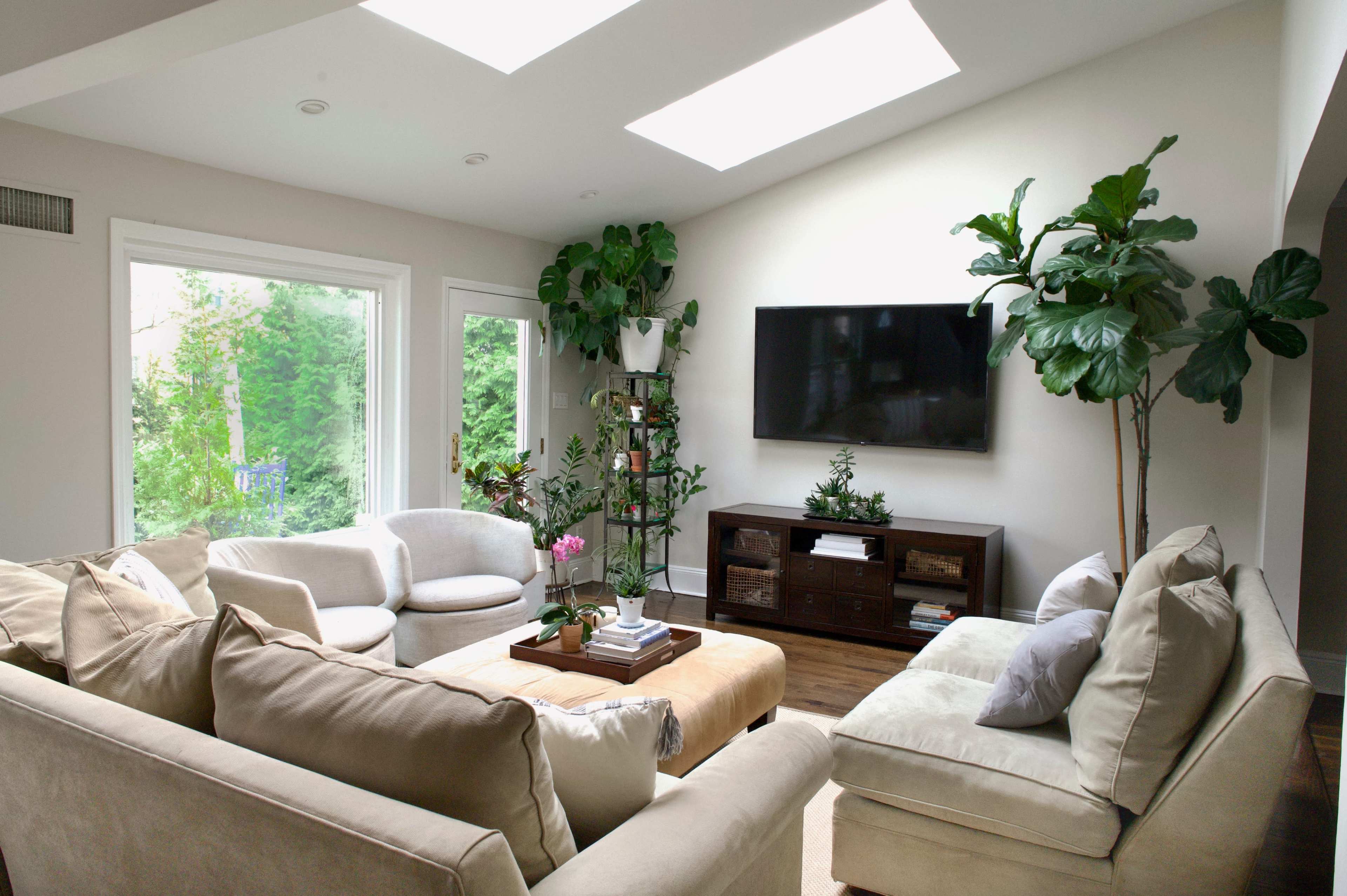 The image shows a well-lit living room with a sectional sofa, a coffee table, a media console, and large windows featuring greenery outside.