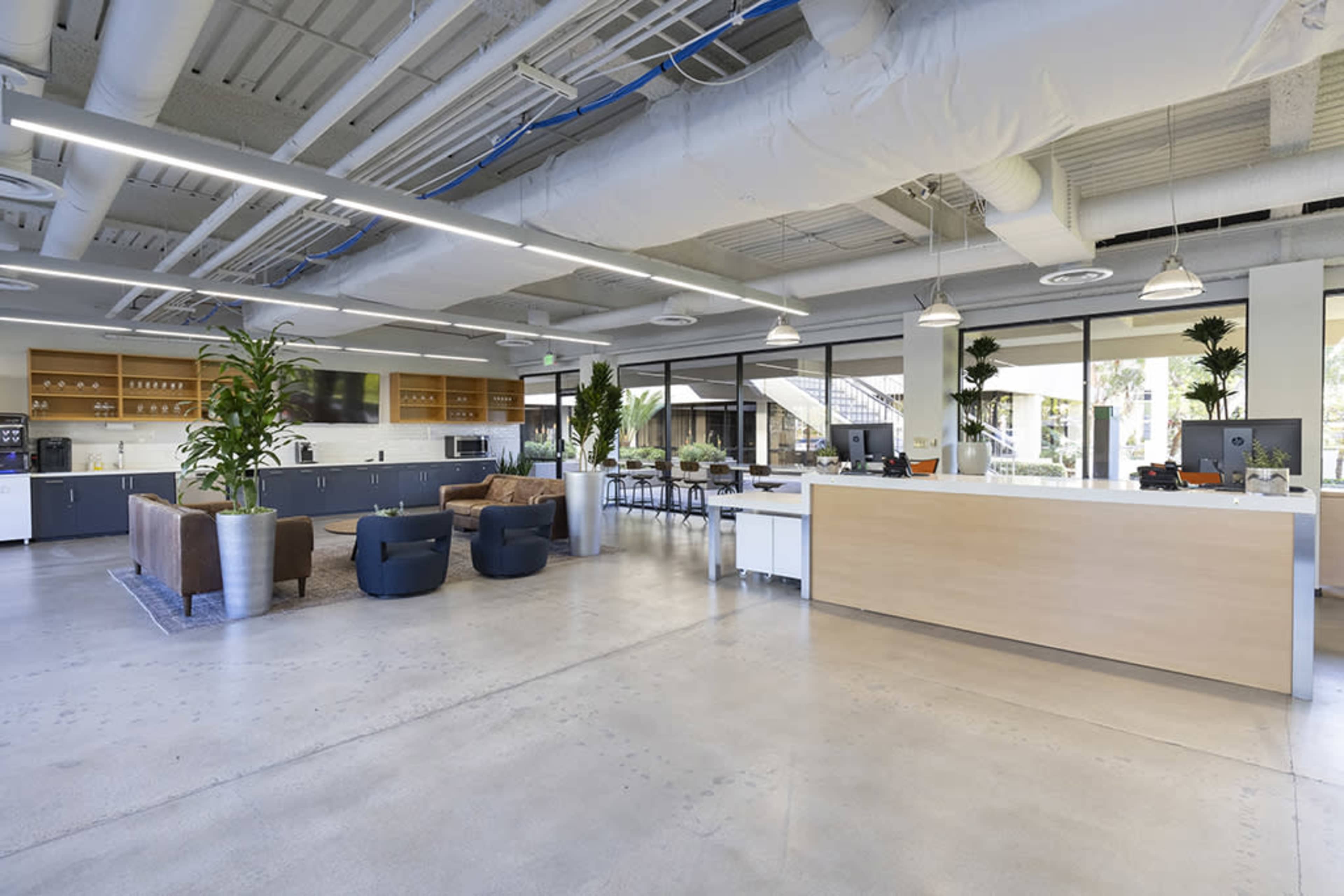 A modern office space featuring seating areas, a reception counter, and a kitchen area with large windows.
