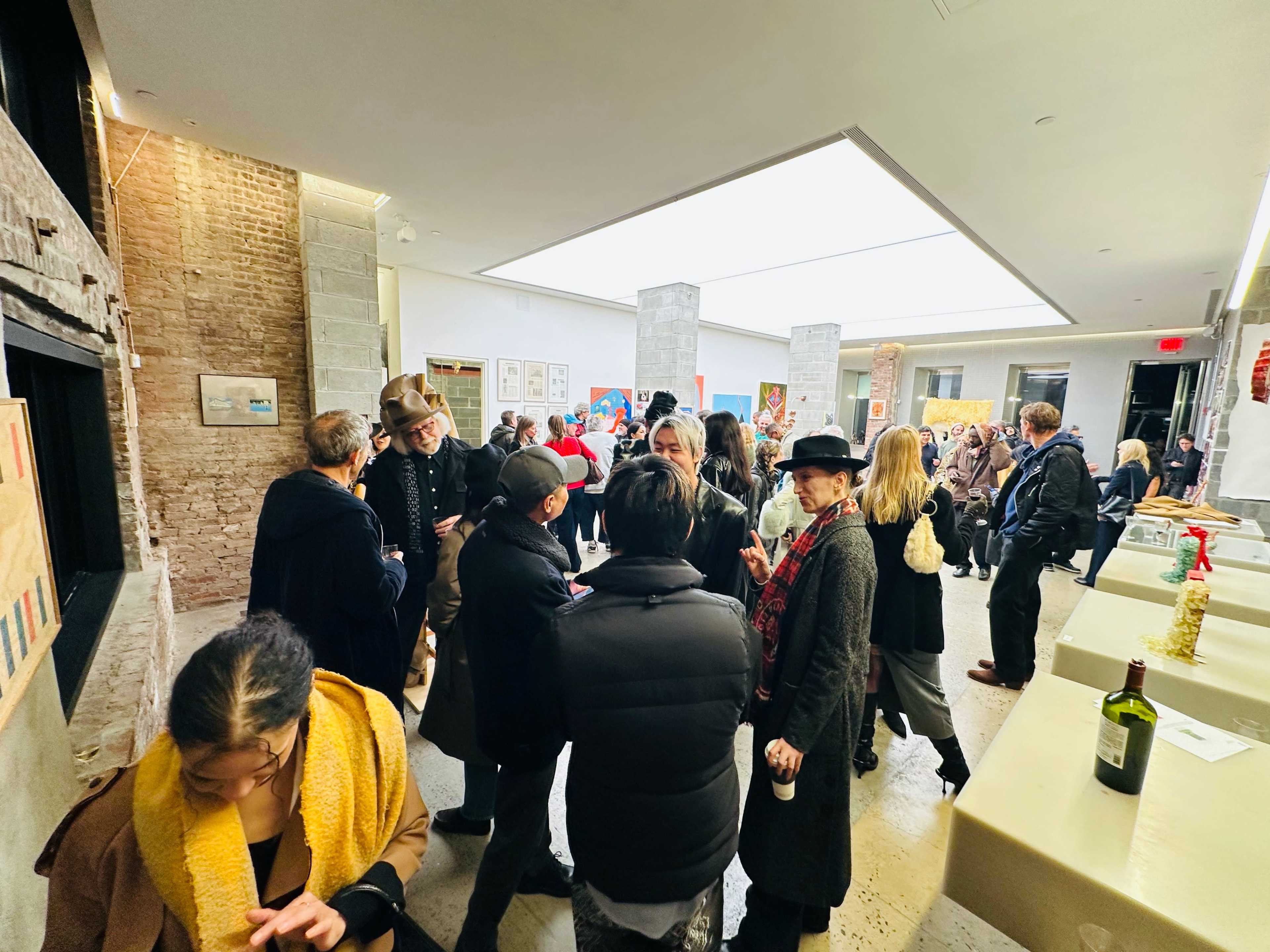 The image shows a crowded art gallery event with people mingling and engaging in conversation, surrounded by artworks on display.