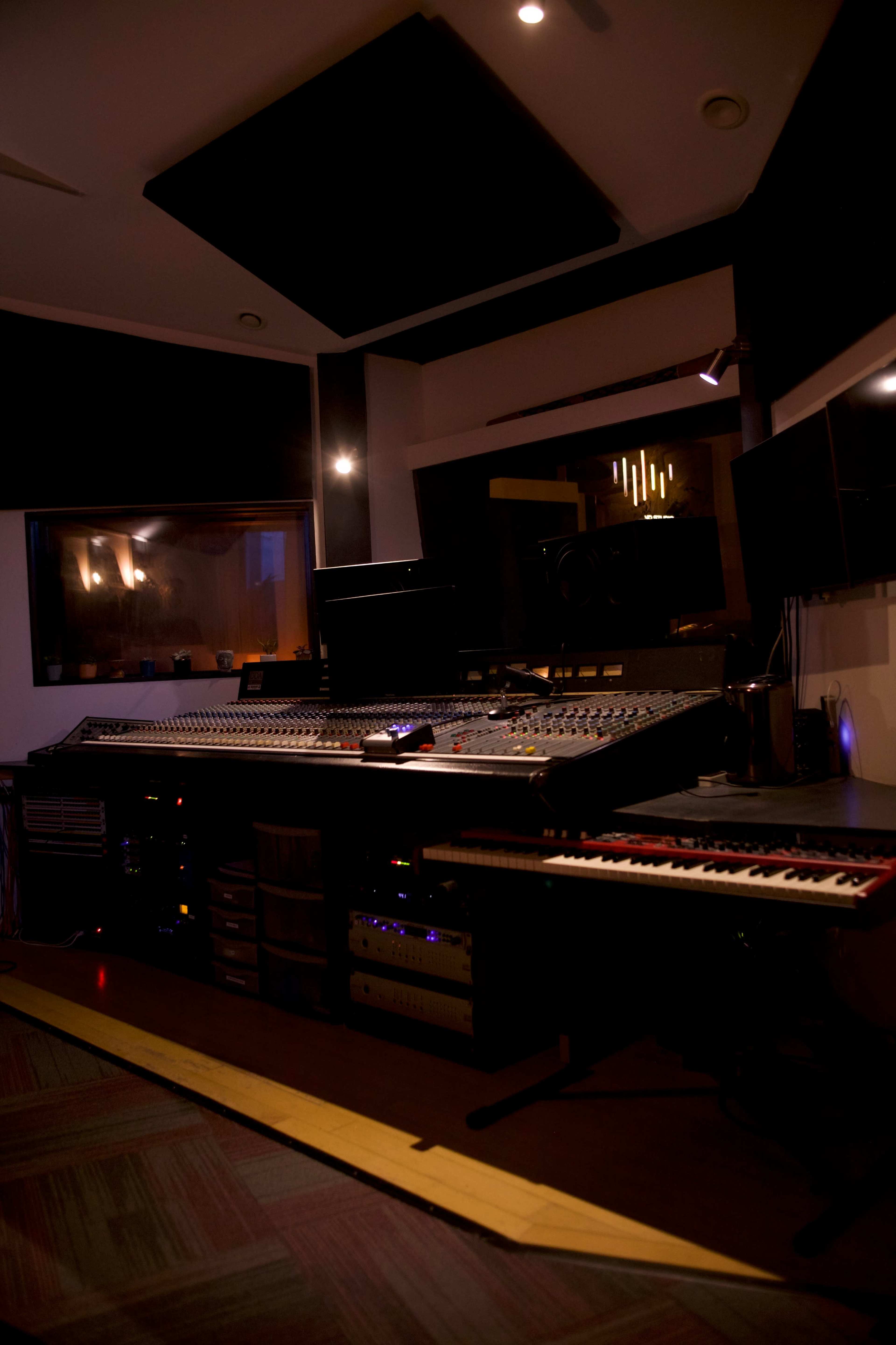 The image shows a music recording studio with a large mixing console, racks of audio equipment, and a keyboard positioned on the floor.