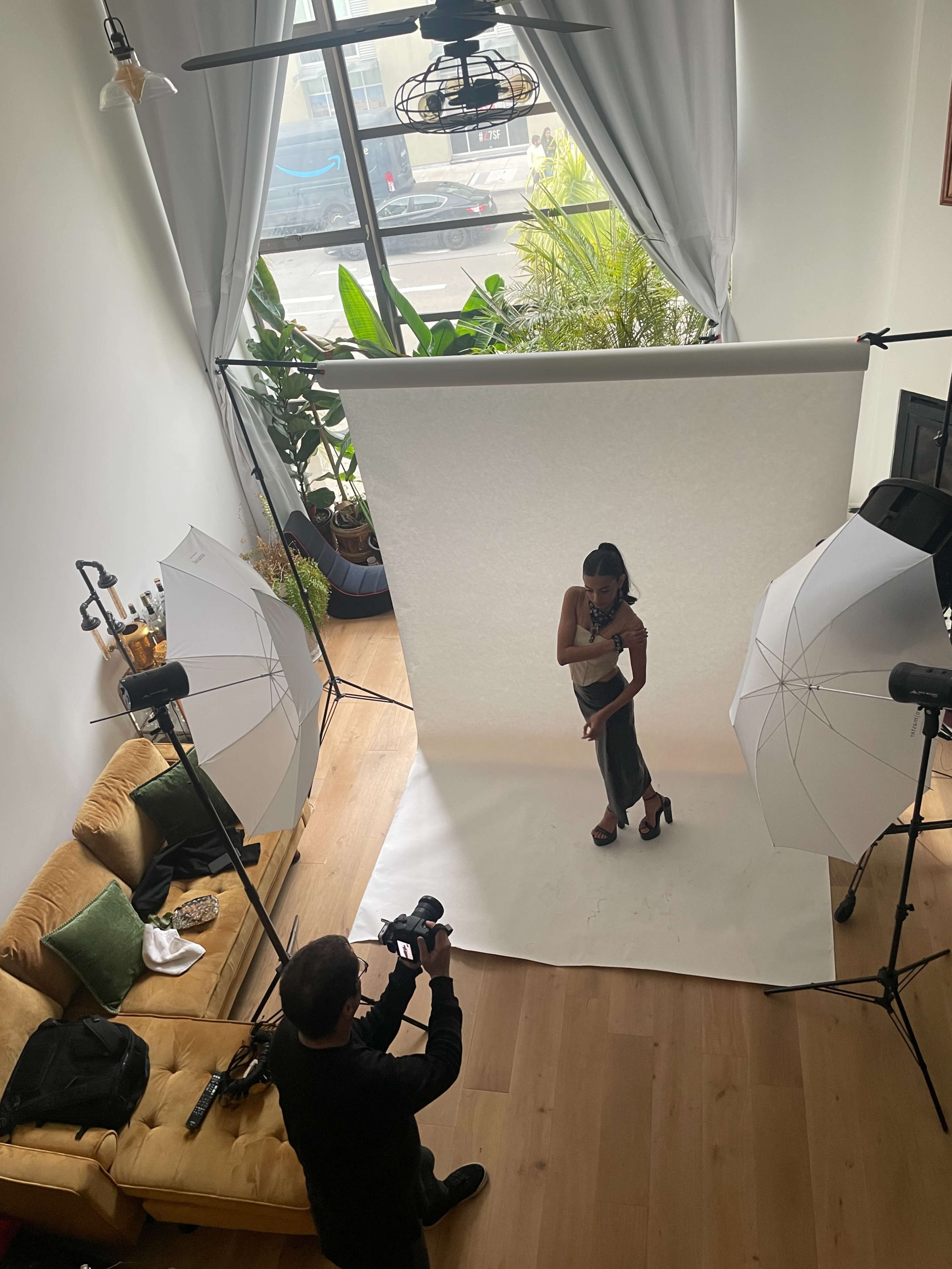 A photographer captures a model's pose in a well-lit indoor space with multiple light setups and a white backdrop.