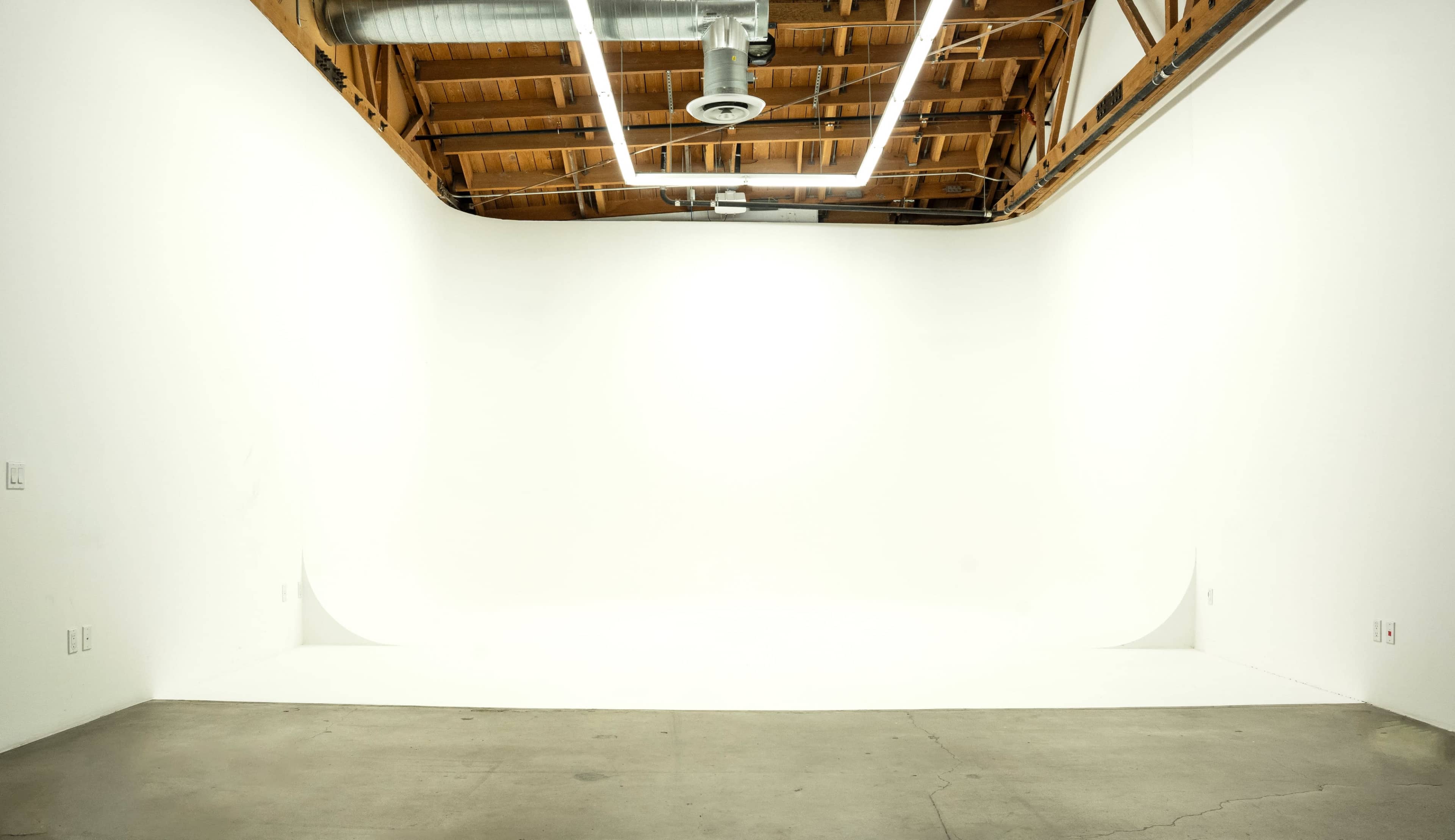 A minimalist photo studio featuring a seamless white backdrop, concrete floor, and exposed wooden beams on the ceiling.