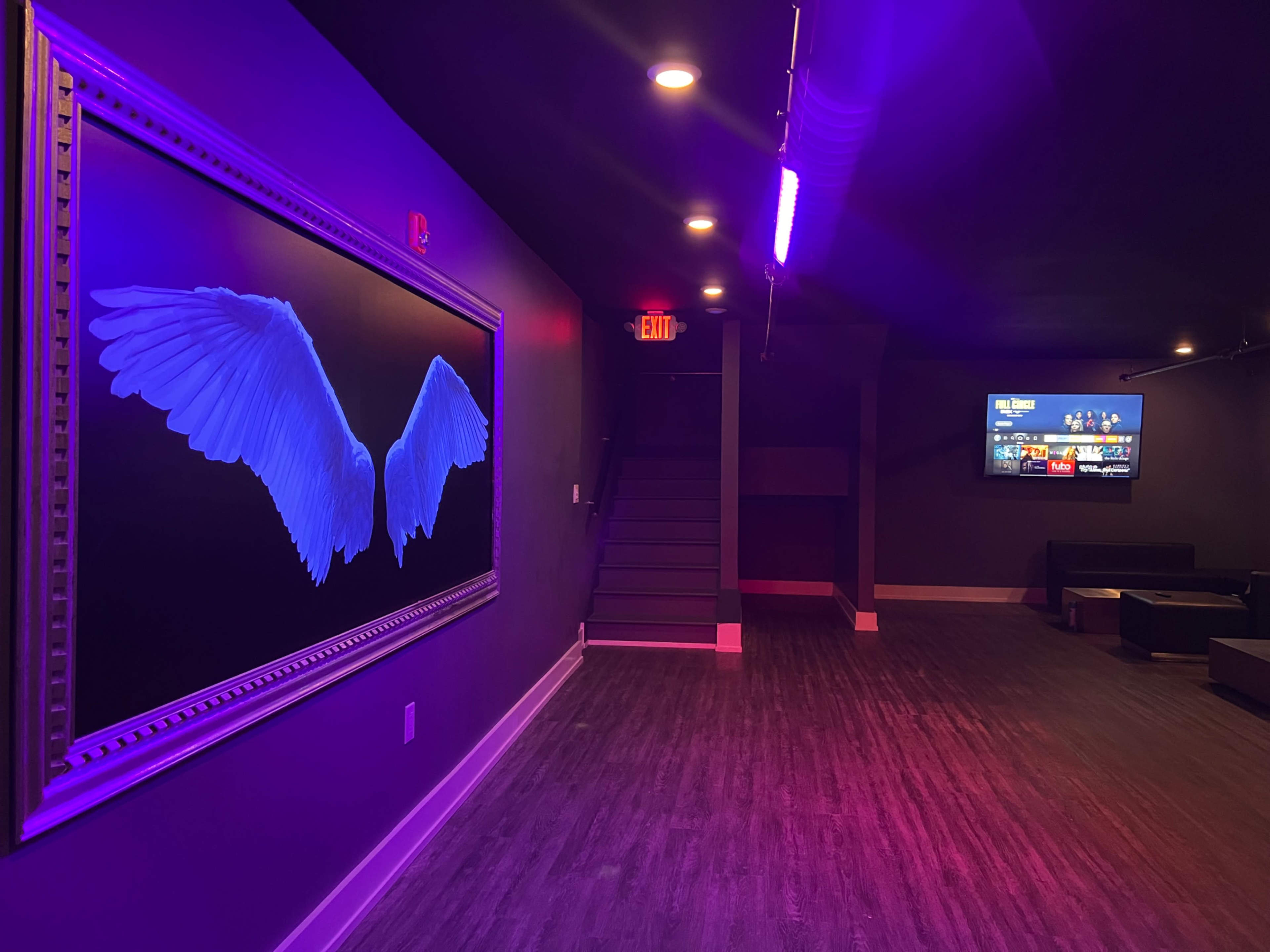 The image shows a dimly lit room featuring a large framed artwork of blue wings on one wall and a screen displaying content on the opposite wall.
