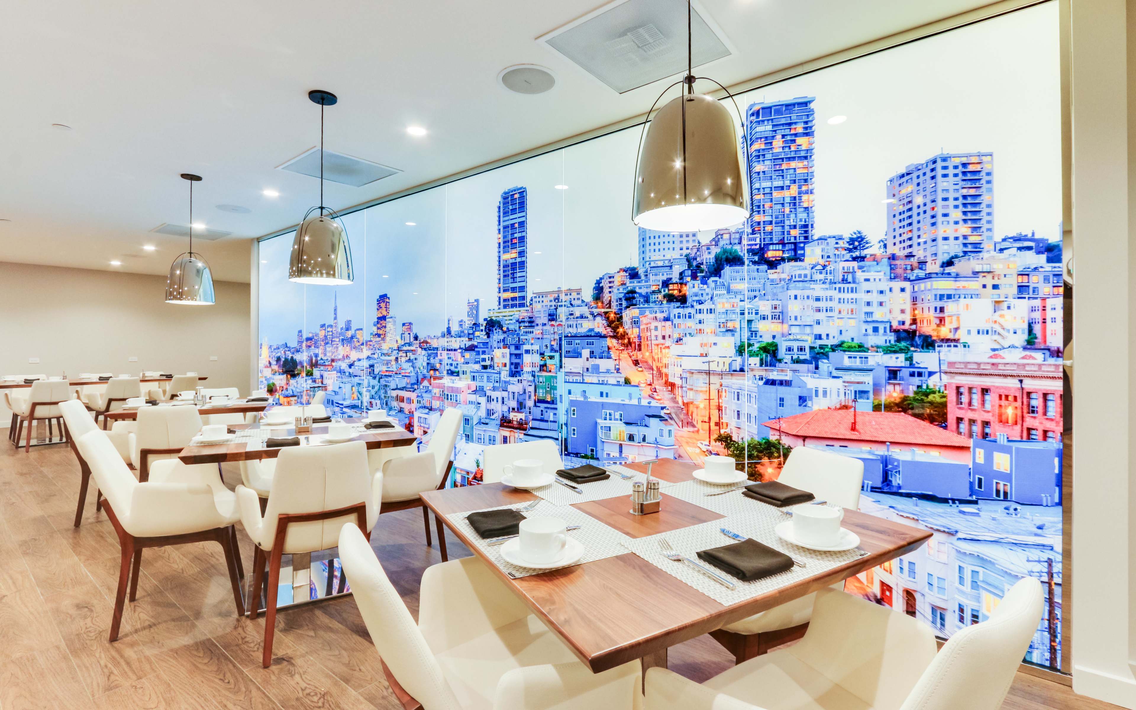 A modern dining area with white chairs and wooden tables, featuring a large mural of a city skyline on the wall.