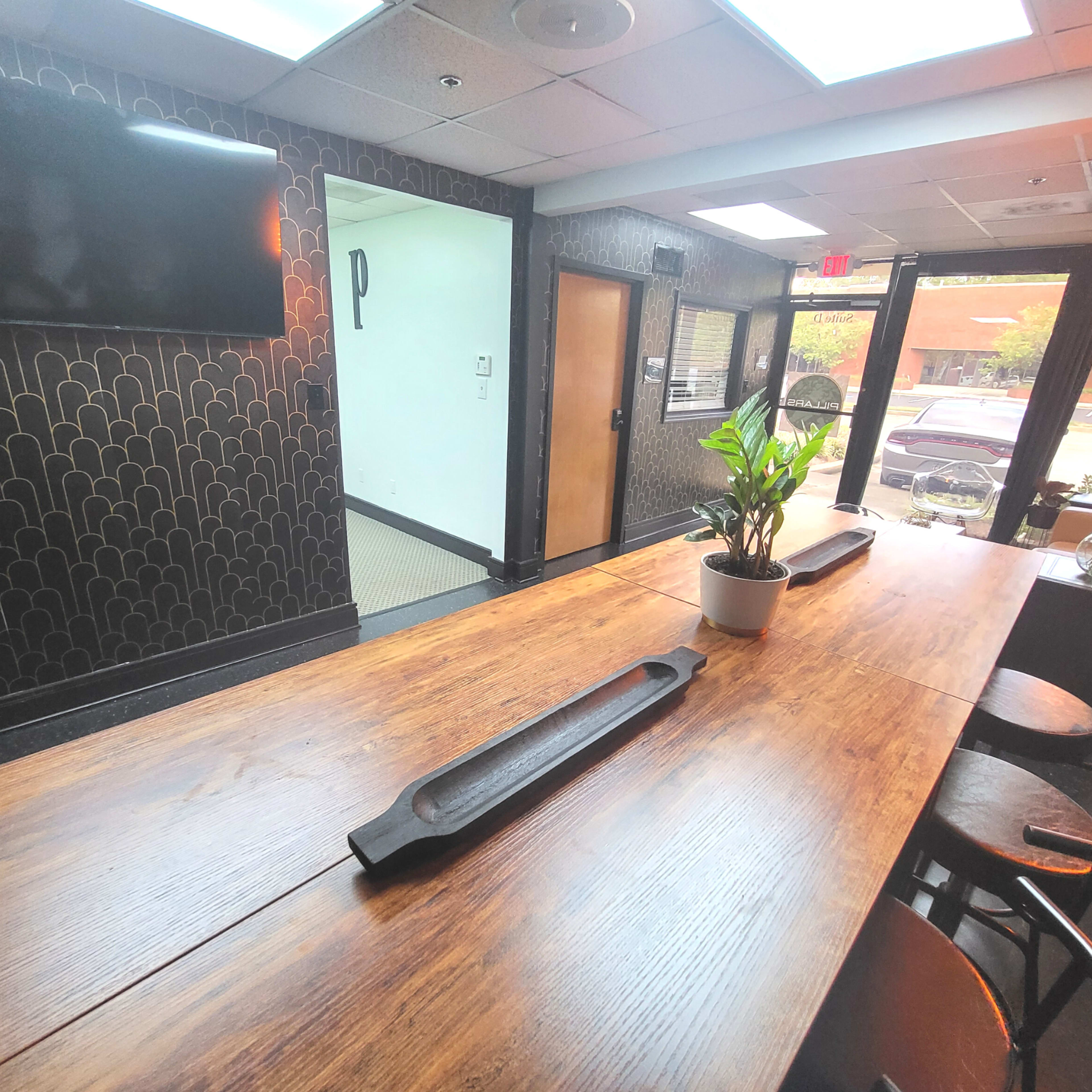 A long wooden table with a decorative centerpiece is set in a well-lit room featuring dark patterned walls and a large window.