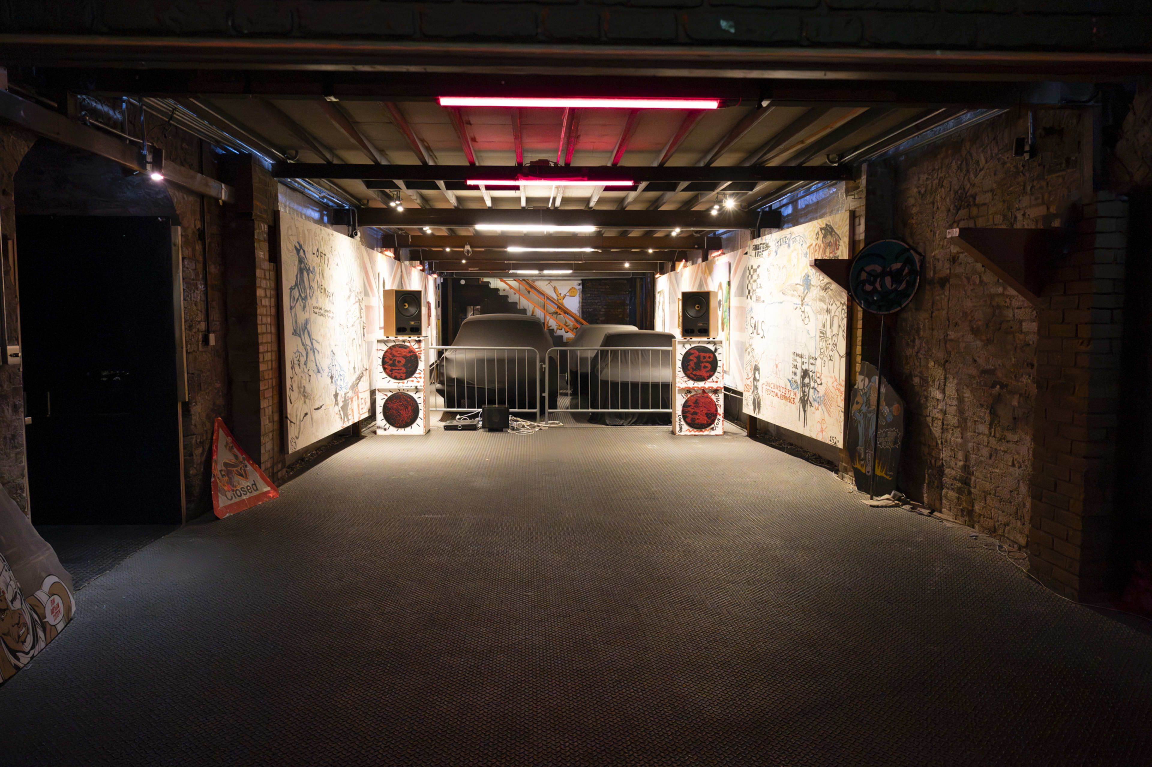 The image shows an industrial space with exposed brick walls, featuring speakers on either side of a covered vehicle and graffiti art on the walls.