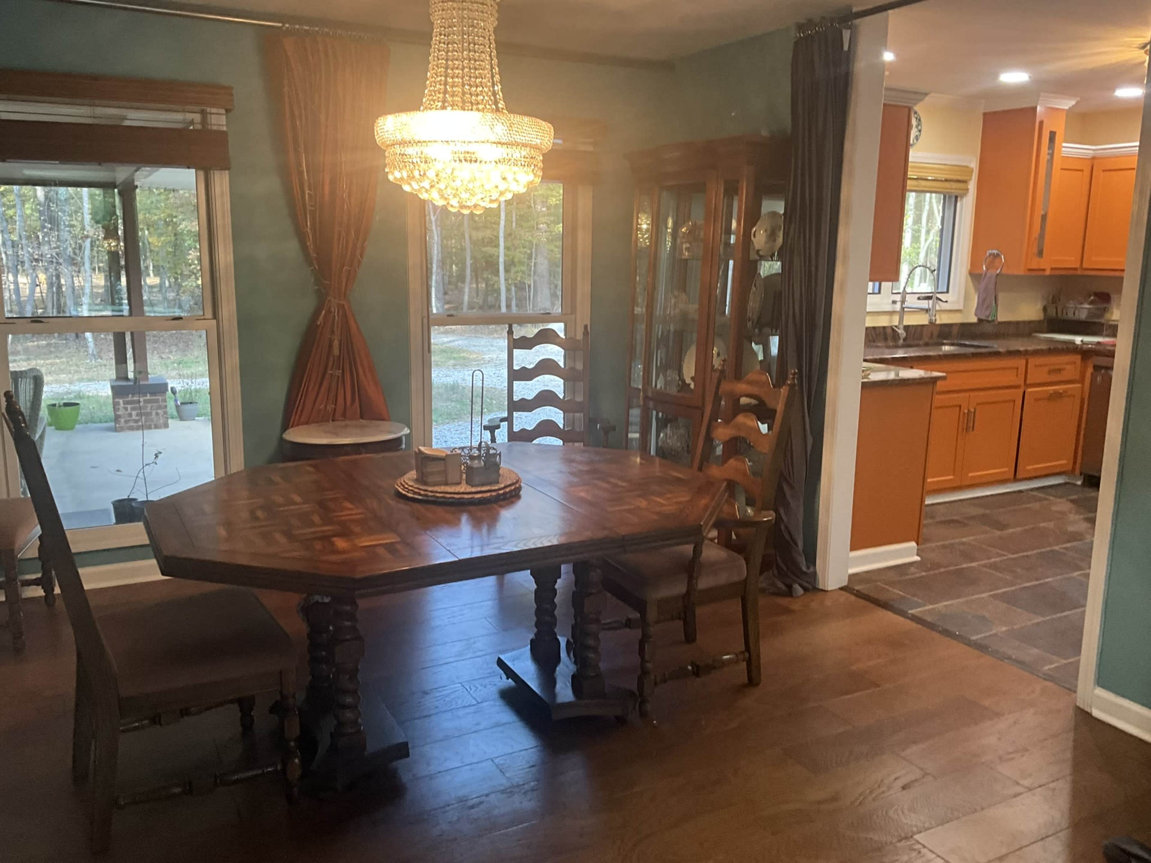 A wooden dining table with chairs is positioned near large windows, with a chandelier overhead and an adjacent kitchen visible in the background.