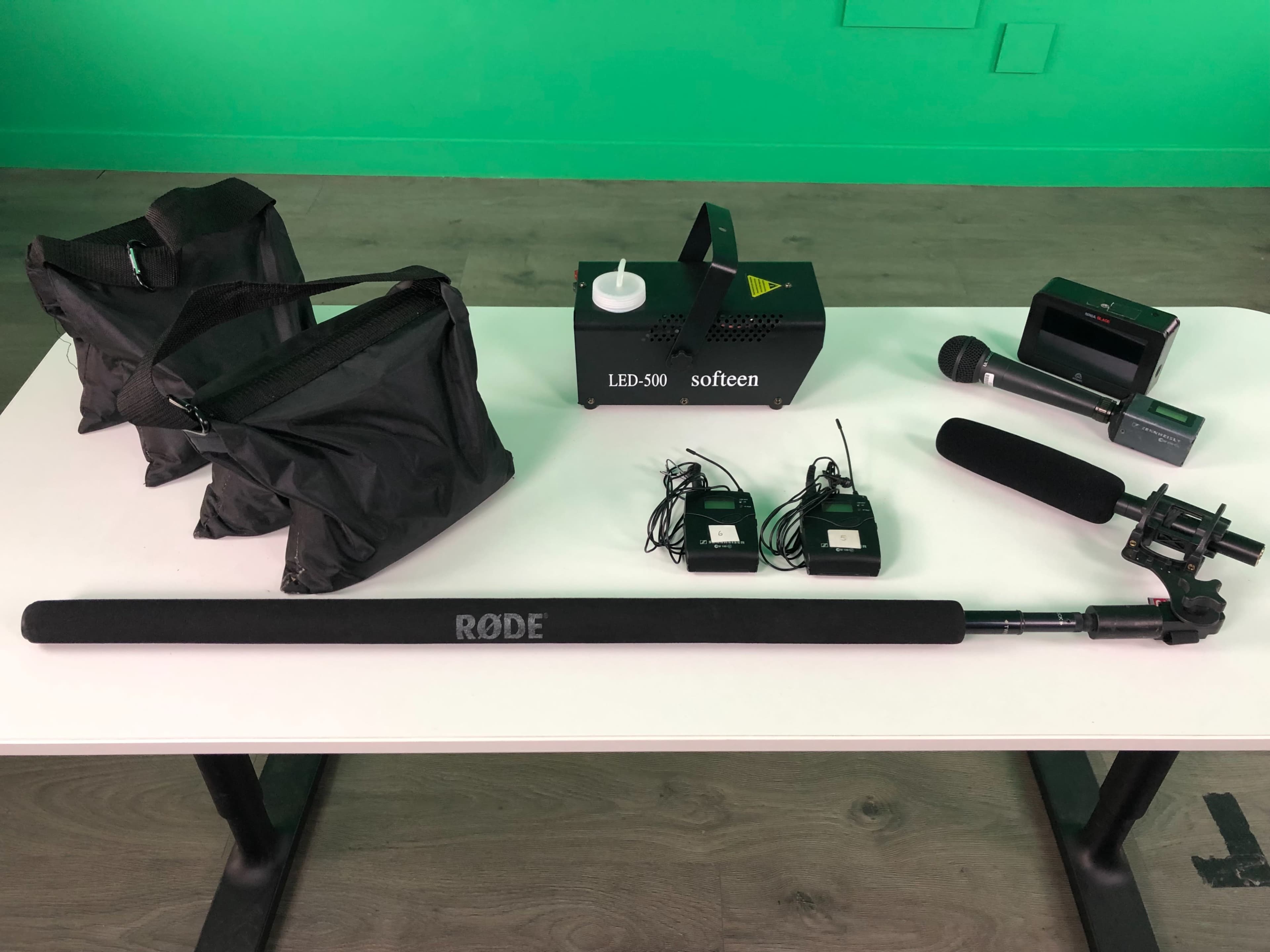 A tabletop arrangement of audio and lighting equipment, including a Rode boom pole, two wireless microphone transmitters, a handheld microphone, a light fixture labeled "LED-500," and two bags.