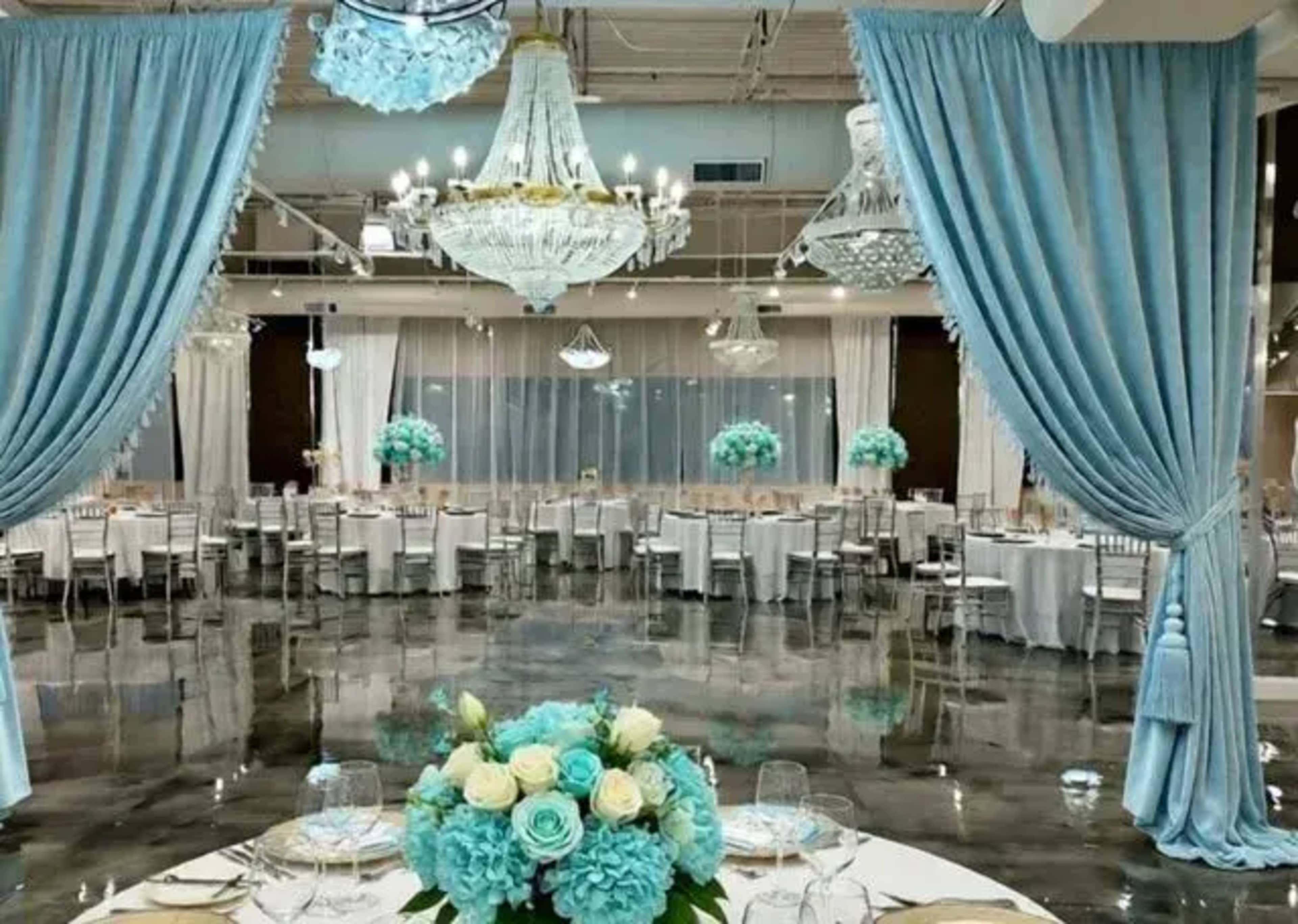 The image shows an elegantly decorated banquet hall with blue drapes, multiple chandeliers, and tables set for an event adorned with floral centerpieces.