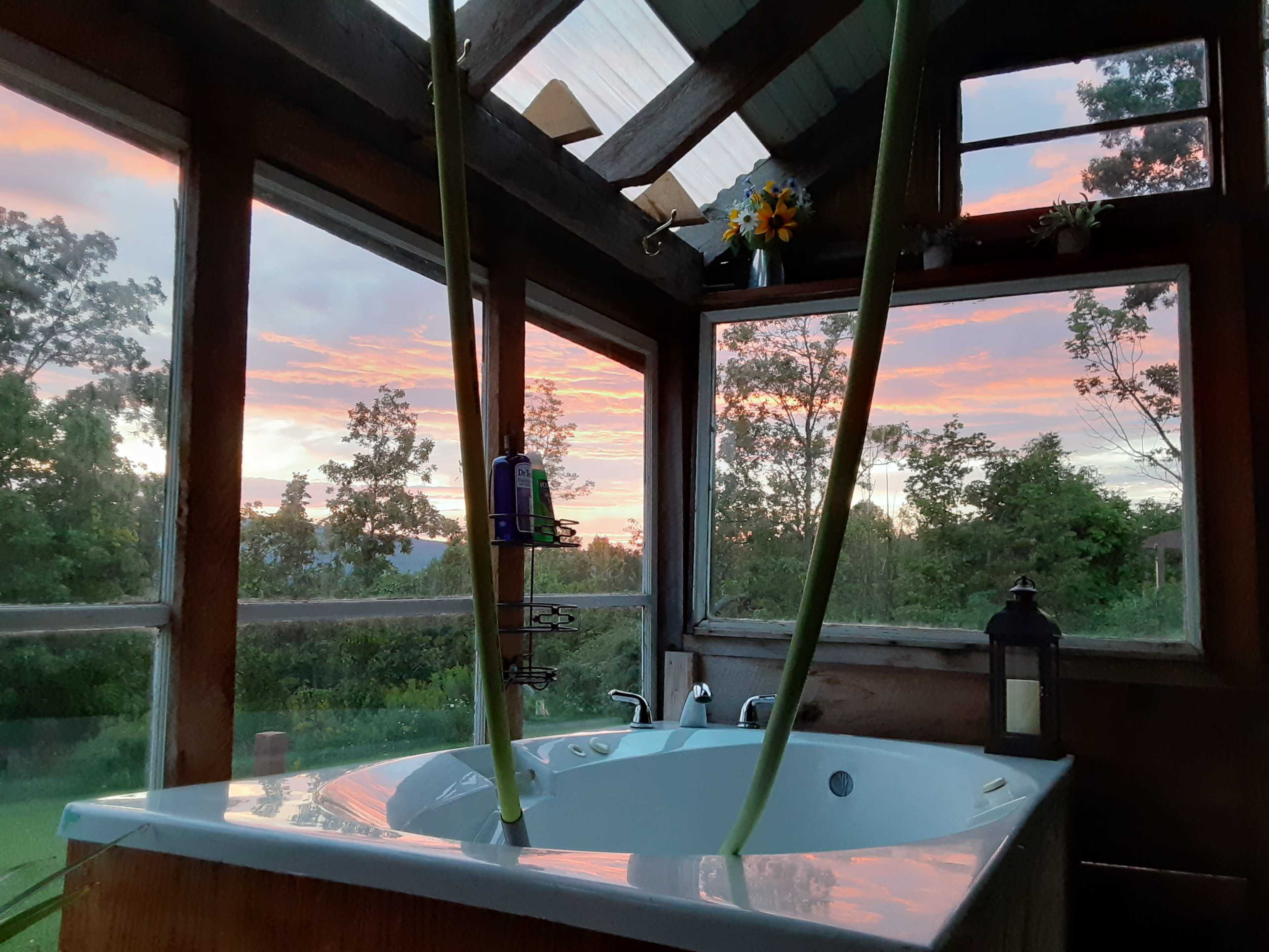 A bathtub inside a wooden structure with large windows, framing a colorful sunset outside.