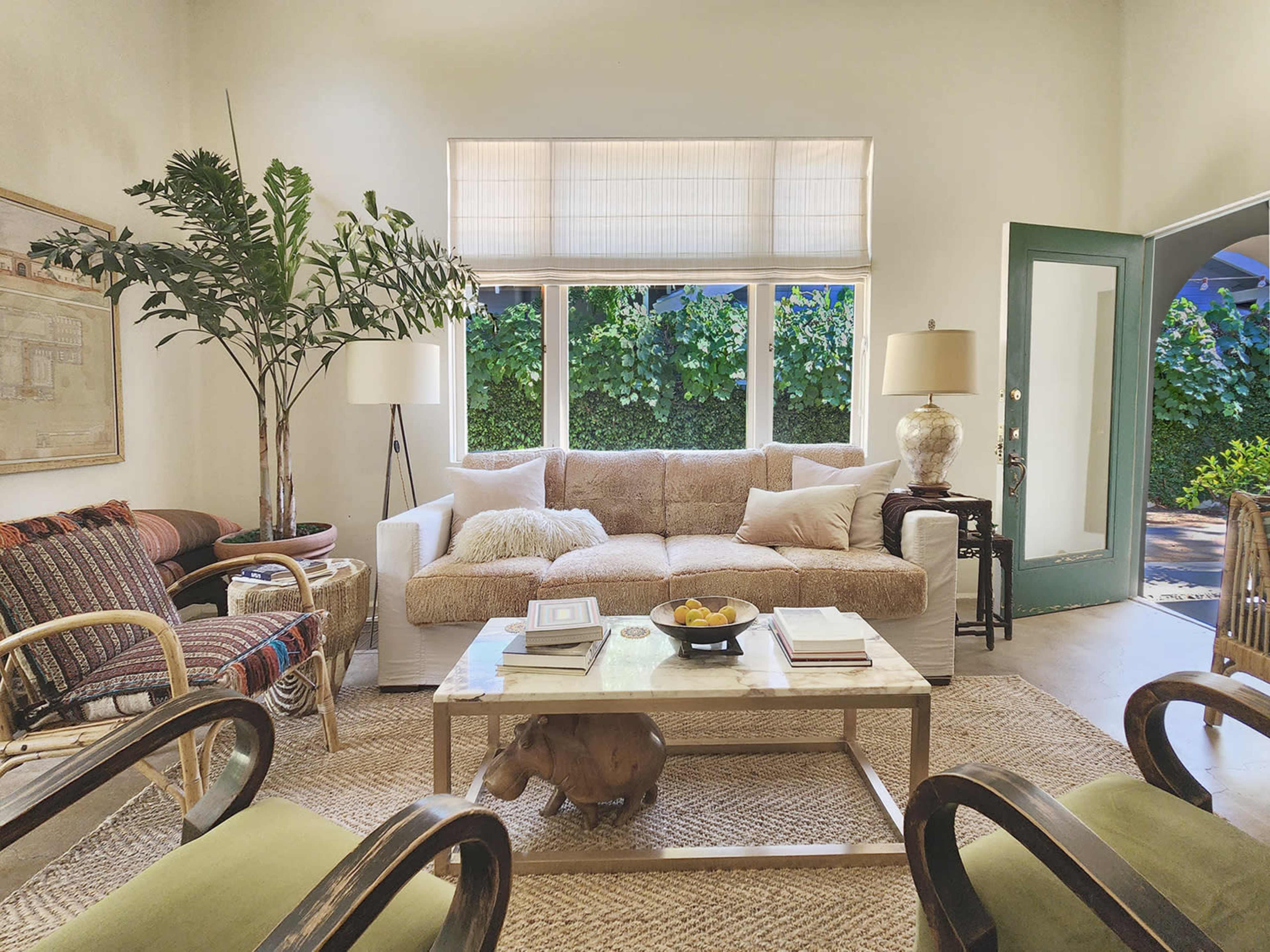 A bright living room features a beige sofa and a coffee table with a bowl of fruit, surrounded by chairs and large windows overlooking greenery.