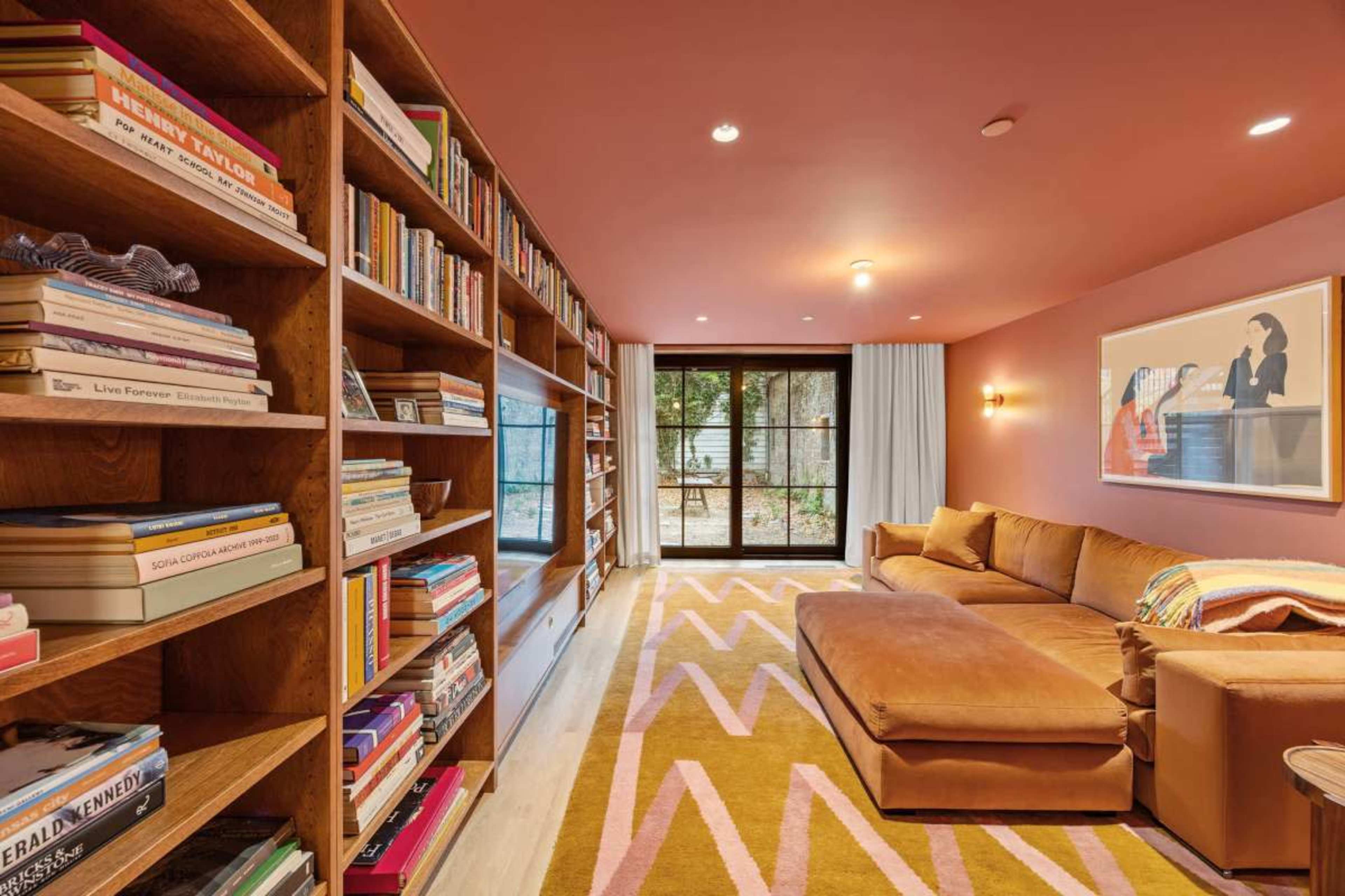 A cozy living room features a large sectional sofa, built-in bookshelves filled with books, and a large window overlooking a garden.