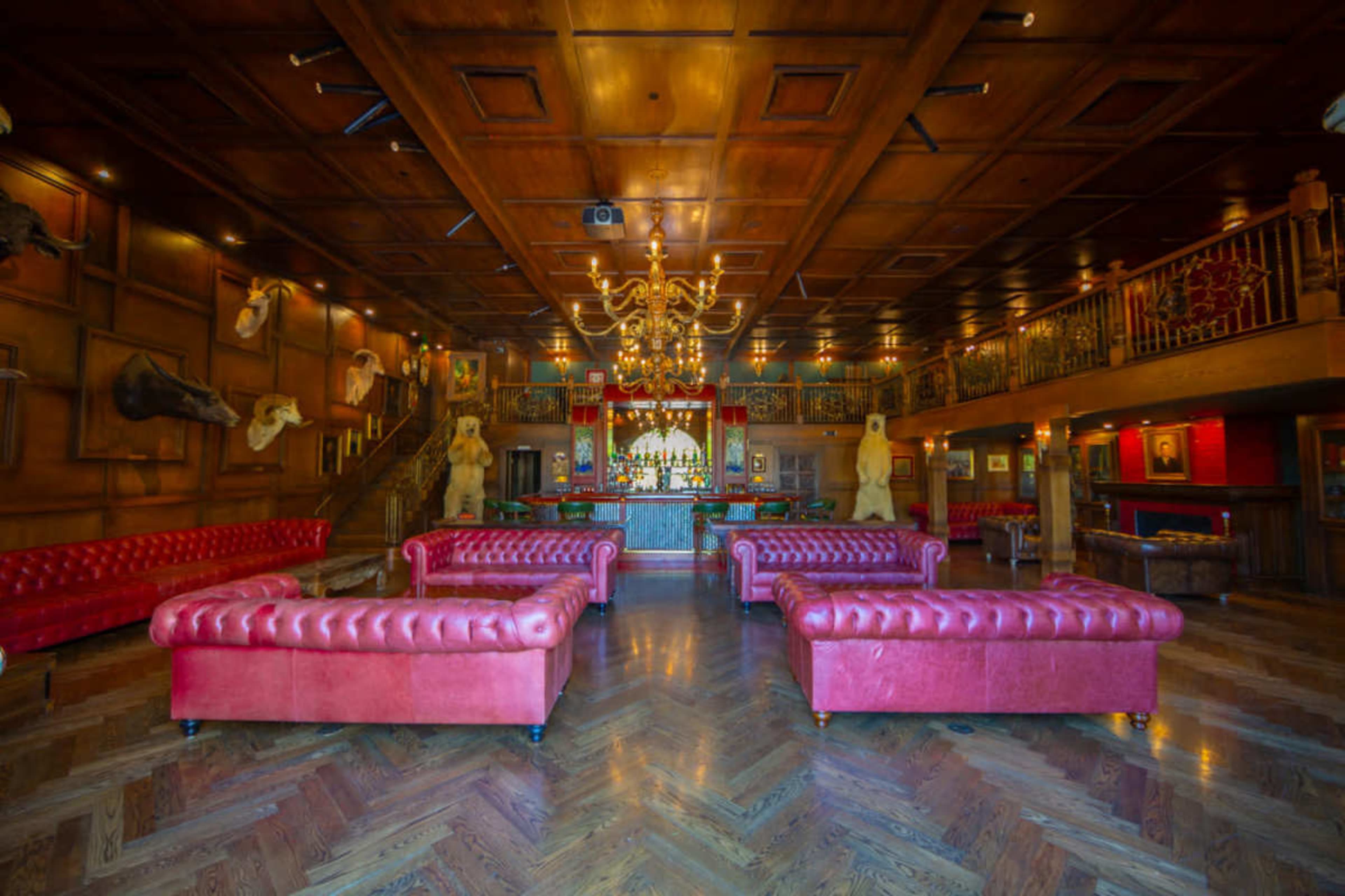 The image shows a spacious room with wooden paneling, featuring red tufted sofas and a bar with decorative elements, including mounted animal heads on the walls.