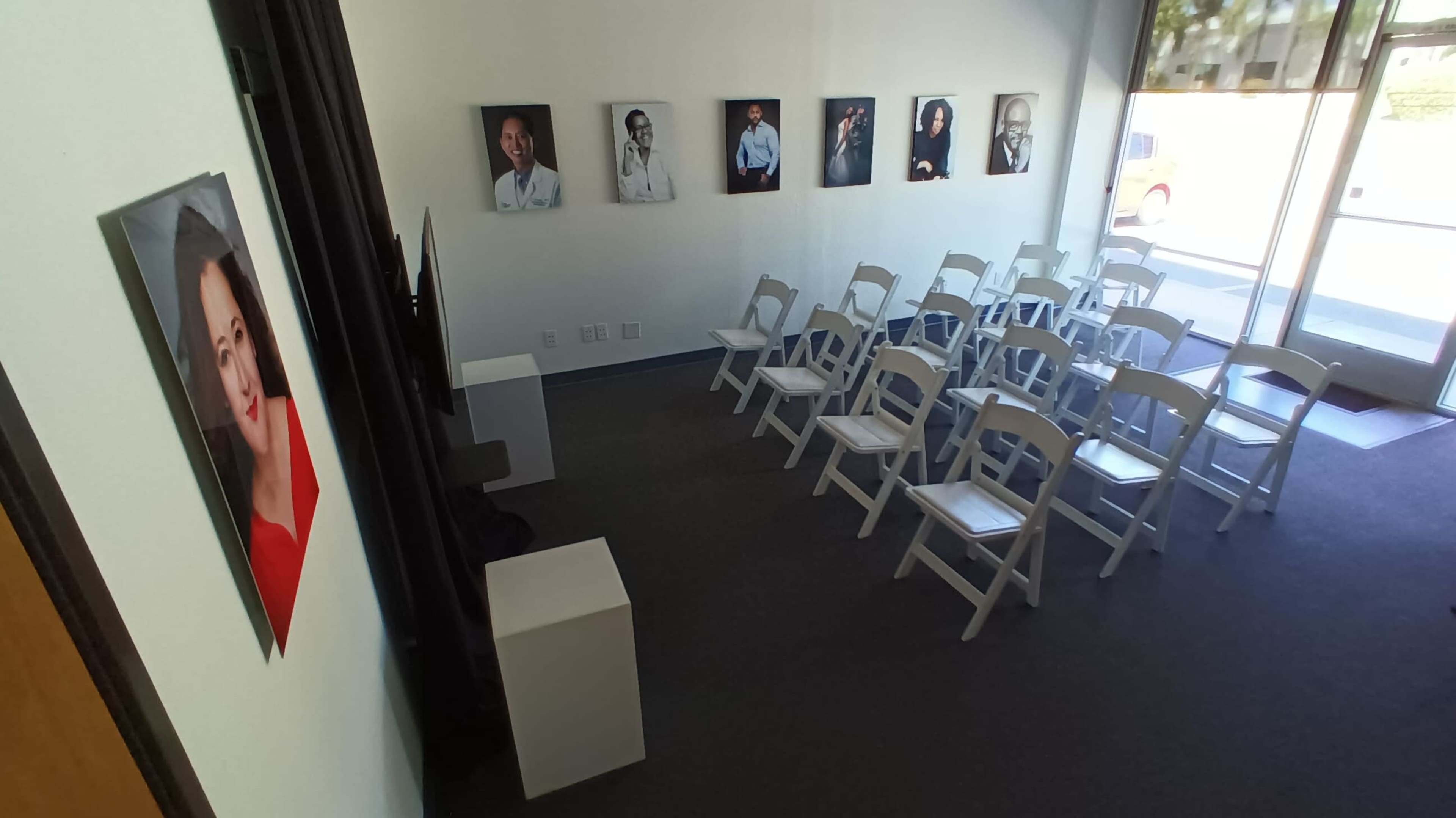 The image shows a room set up with white chairs facing a blank wall, adorned with framed portraits on the adjacent walls.