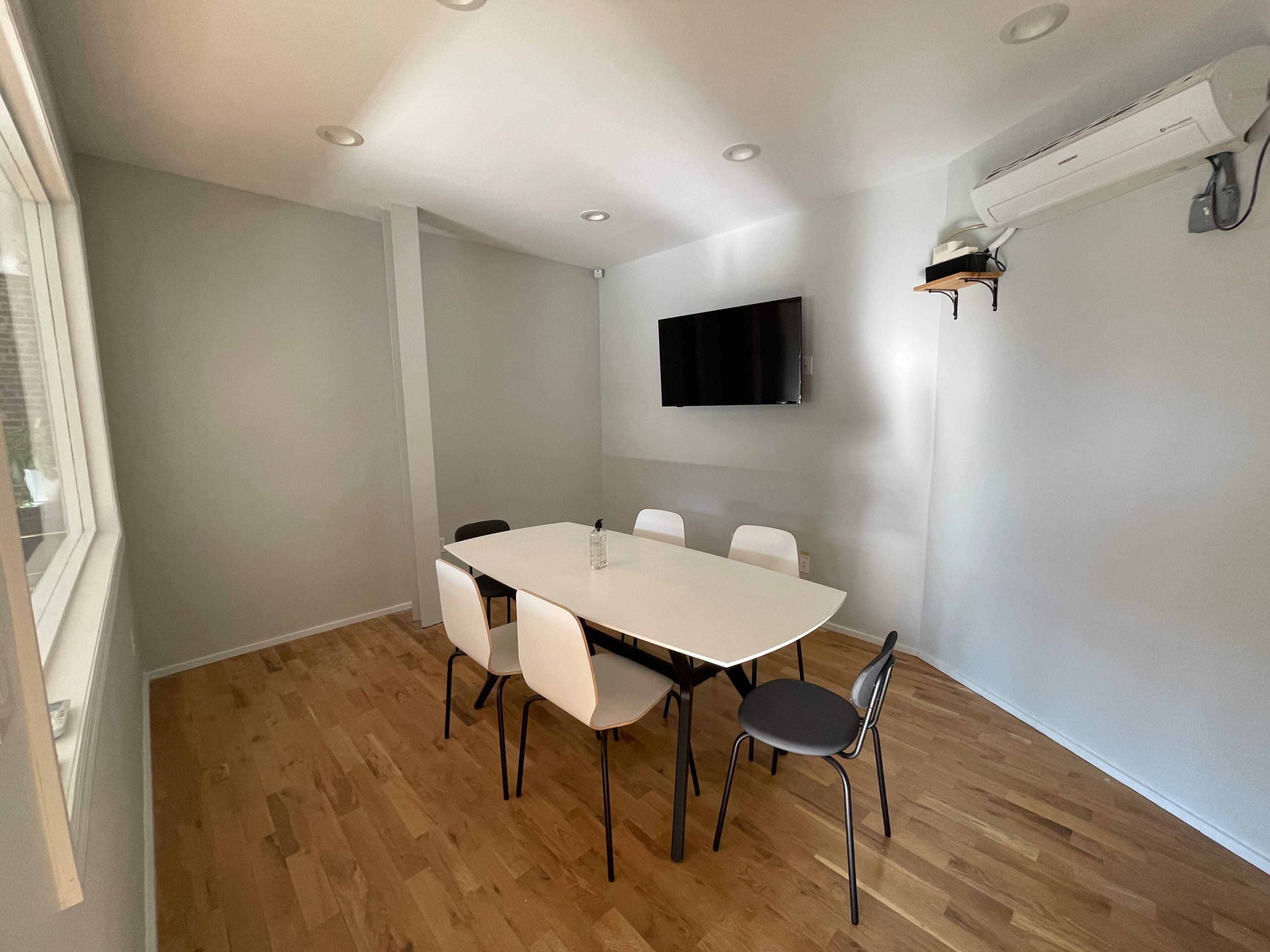 A light-colored meeting room with a large table surrounded by six chairs and a wall-mounted television.