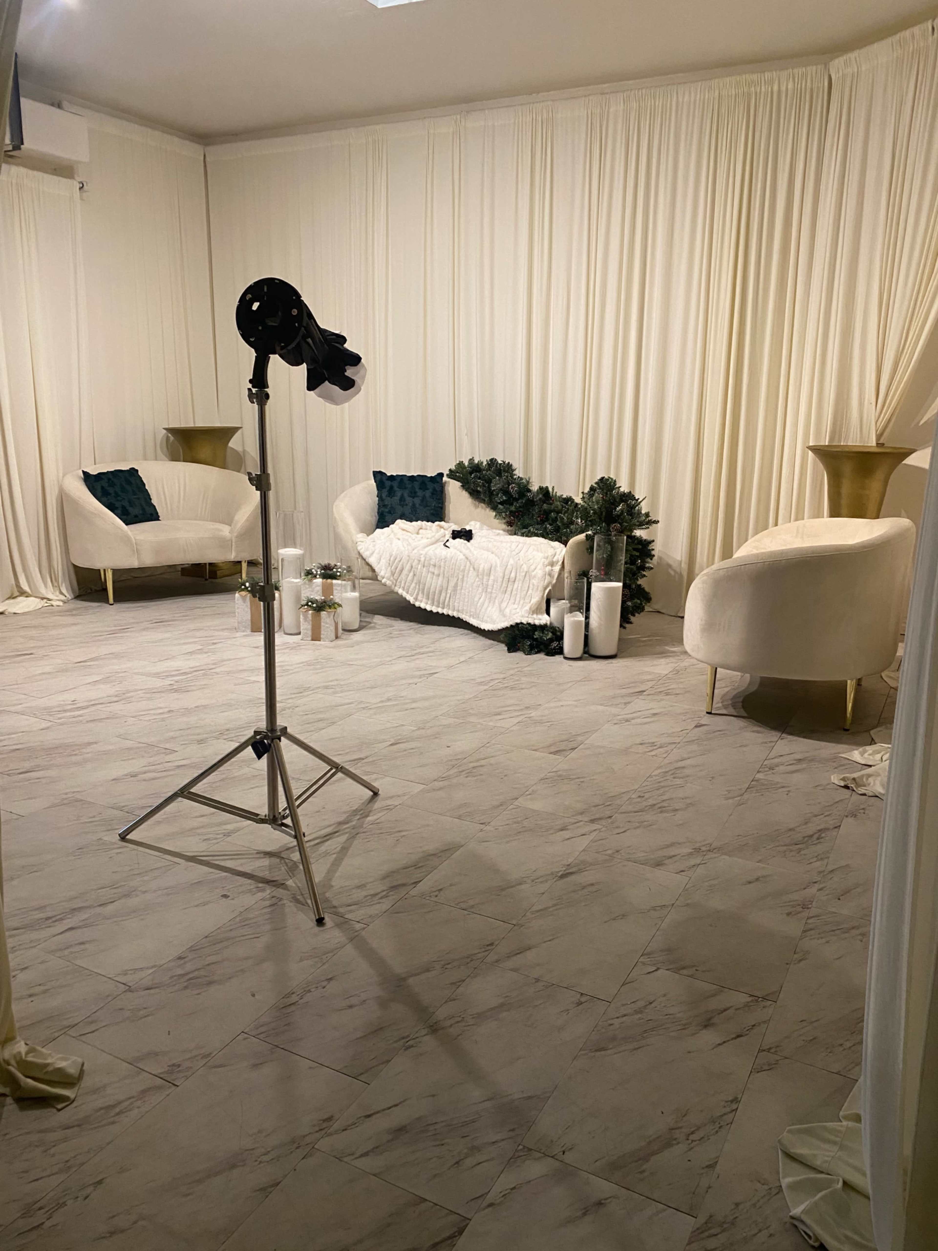 The image shows a minimalist studio setup with two light-colored chairs, a white blanket draped over one, and decorative greenery arranged on the floor against a backdrop of white curtains.