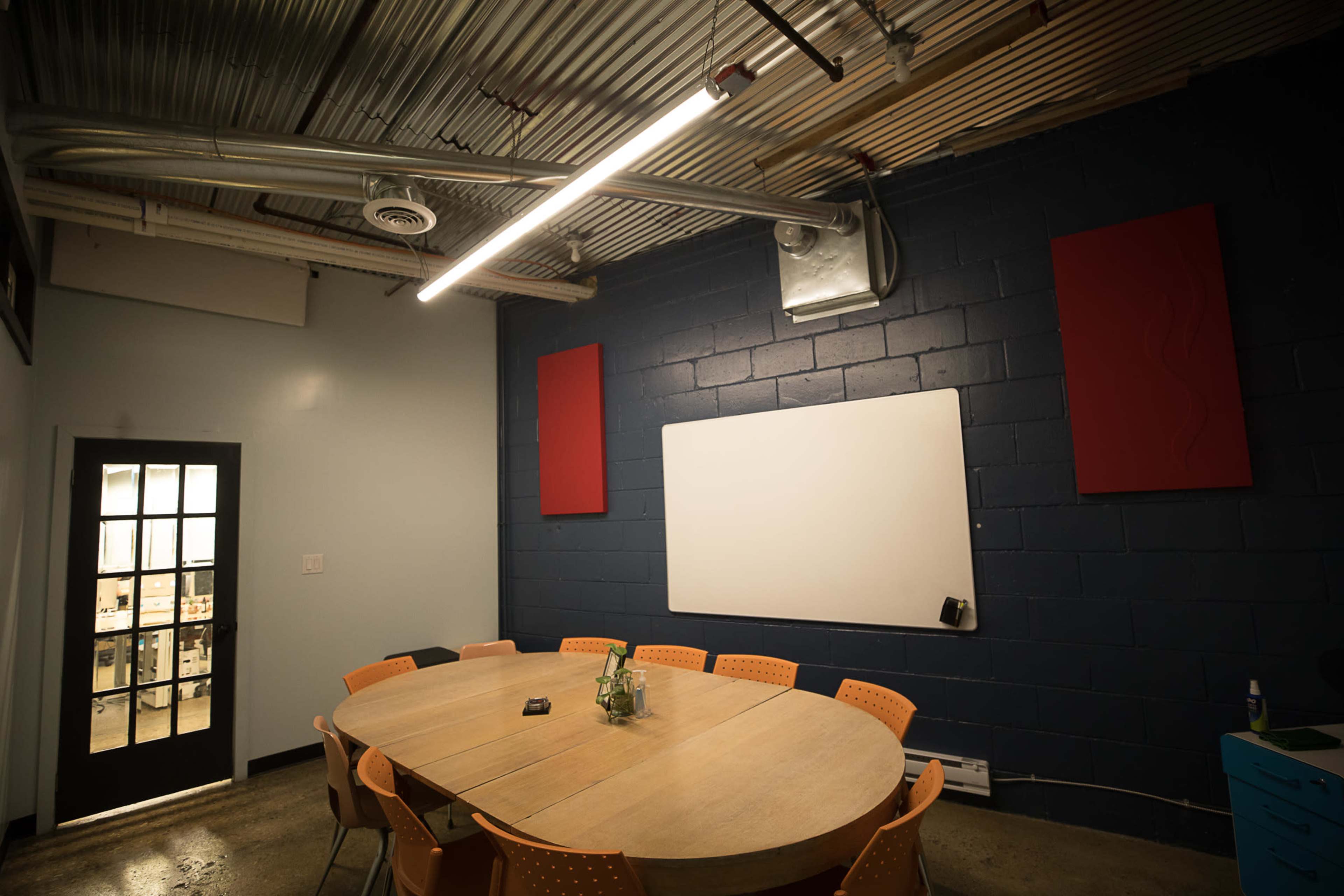 A conference room features a round wooden table surrounded by orange chairs, a whiteboard on the wall, and red wall panels.