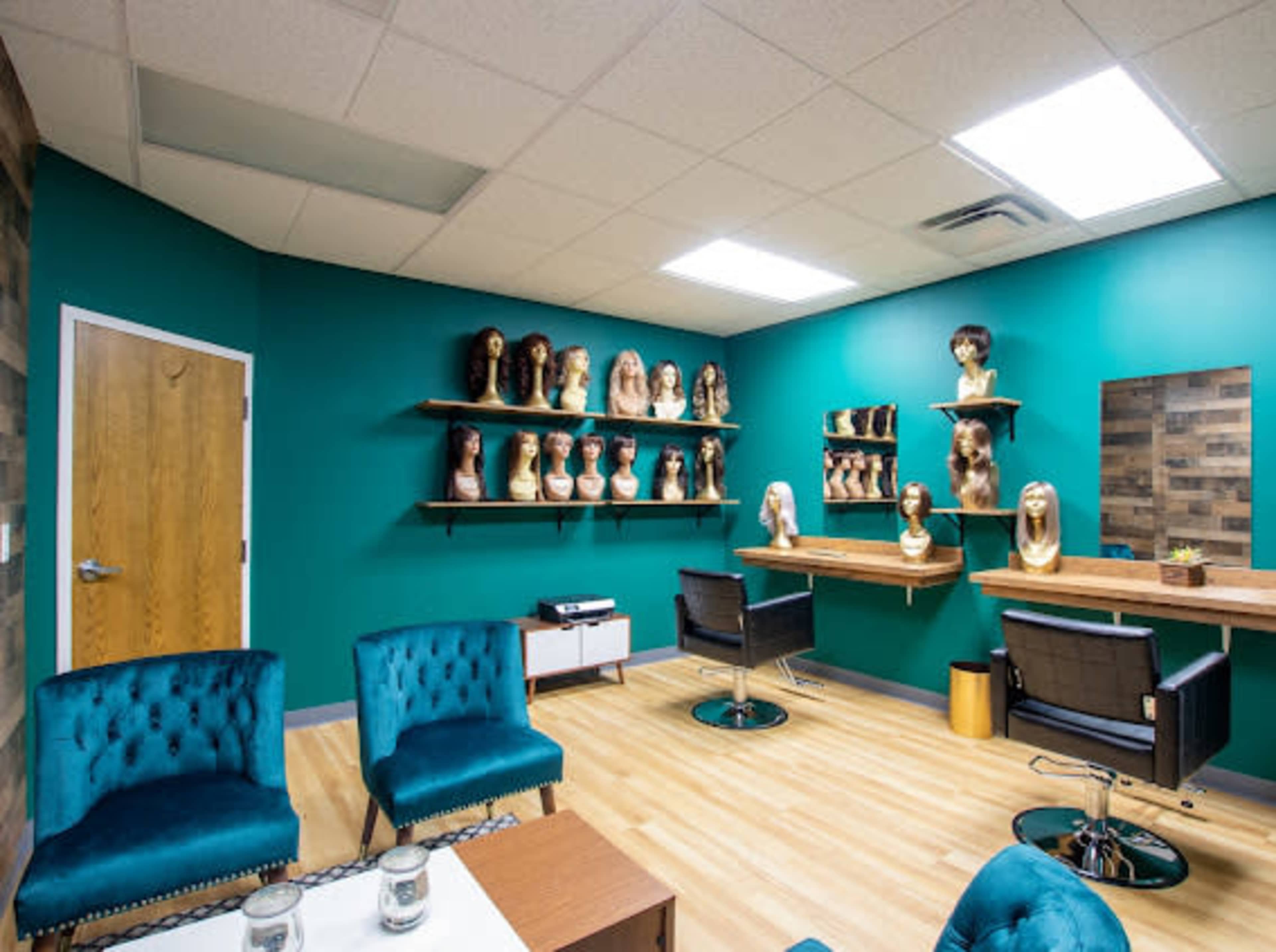 A brightly colored salon interior with teal walls, a display of wigs on shelves, and modern furniture including chairs and a table.