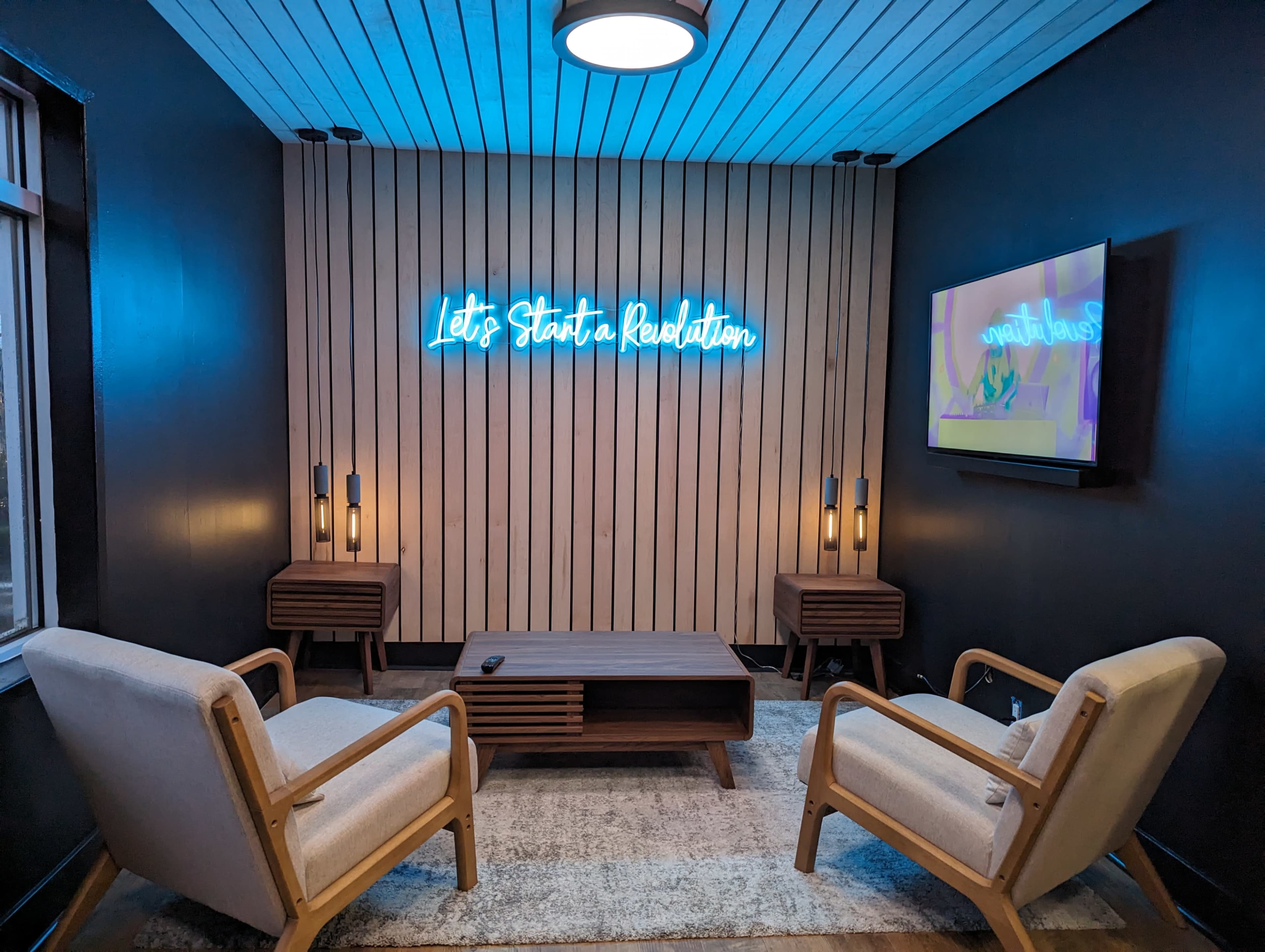 A small, modern lounge area with two wooden chairs, a coffee table, and a neon sign on the wall that reads "Let's Start a Revolution."